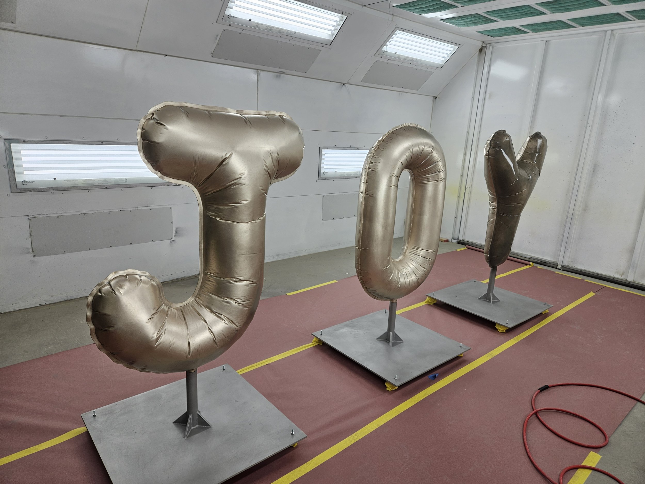 Large metallic balloons shaped like the letters J, O, and Y in a white room with a red floor mat, yellow lines, and ceiling windows.