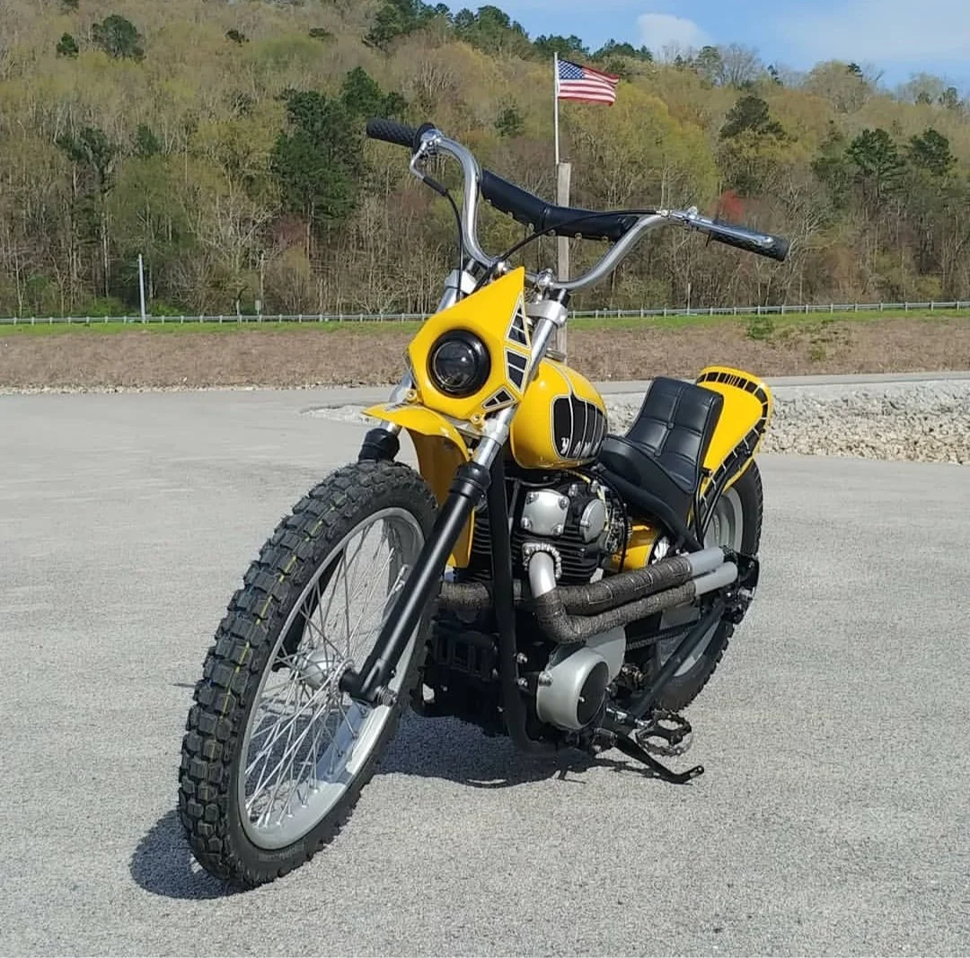 A custom yellow and black motorized tricycle with wide tires, positioned on a paved area, with a background of a hillside, trees, and an American flag.