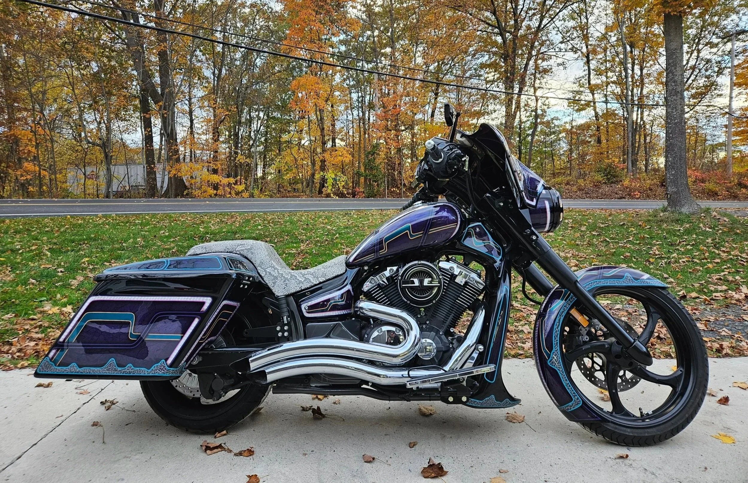 A custom black motorcycle with blue and purple accent details parked on a sidewalk with fallen leaves, against a background of trees with orange, yellow, and green autumn foliage and a road.