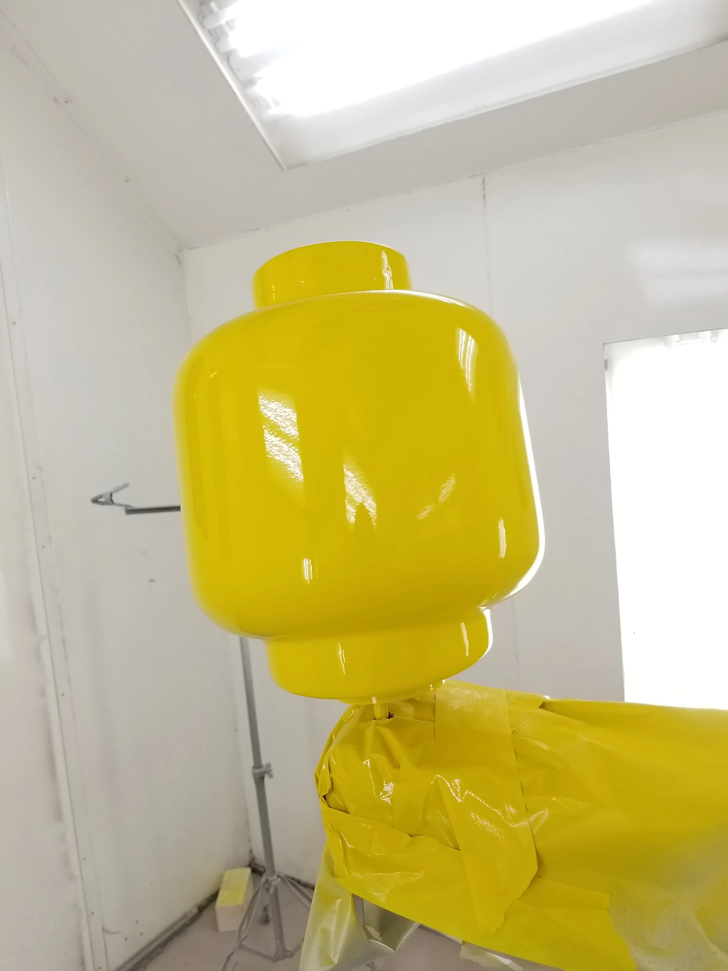 A large yellow LEGO head sculpture on a stand in a white room, with fluorescent lighting overhead.