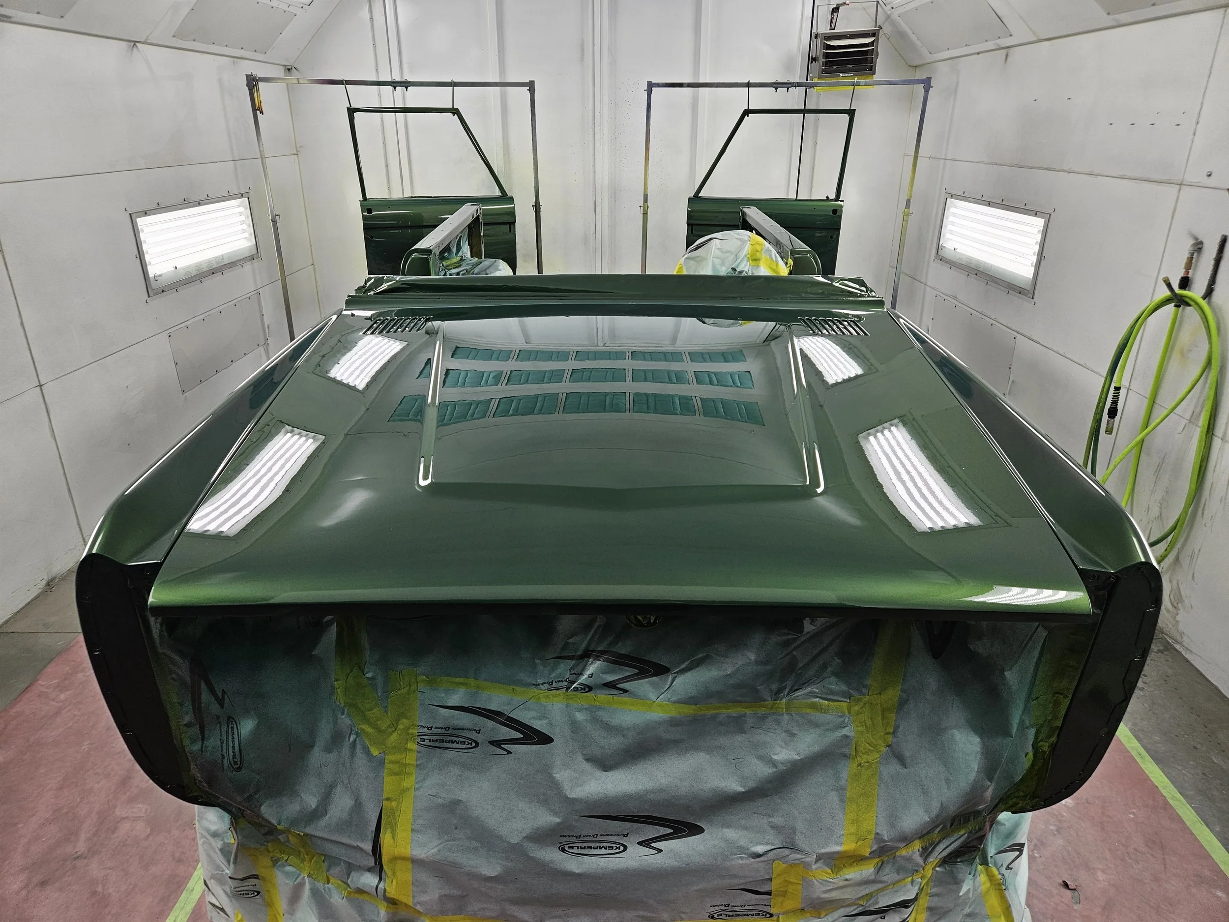 A green car in a paint booth with the front covered in masking paper and taped for painting.