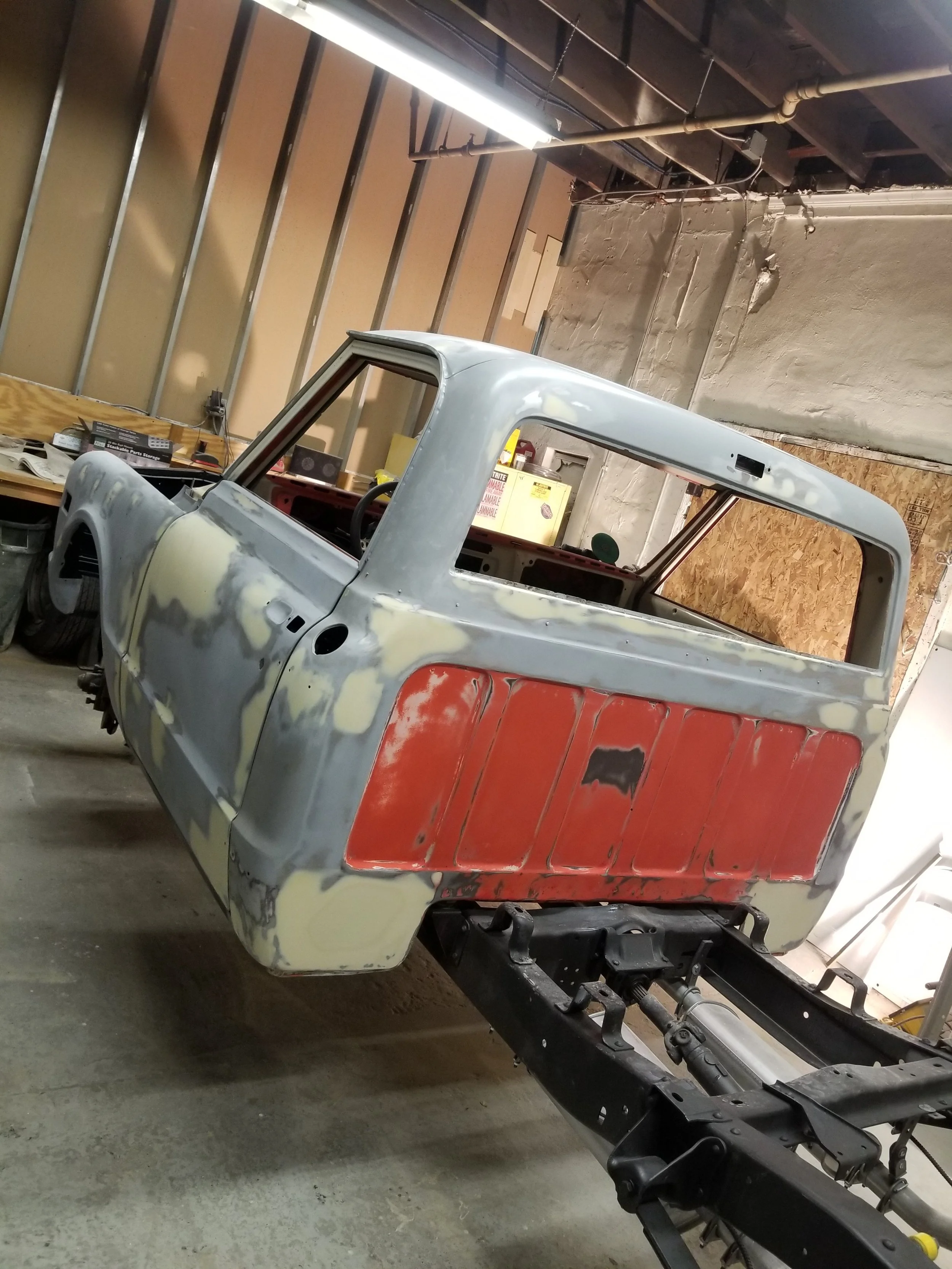 A partially restored or repaired pickup truck body in a workshop, with no wheels or engine, showing primer, rust, and patches of body filler.