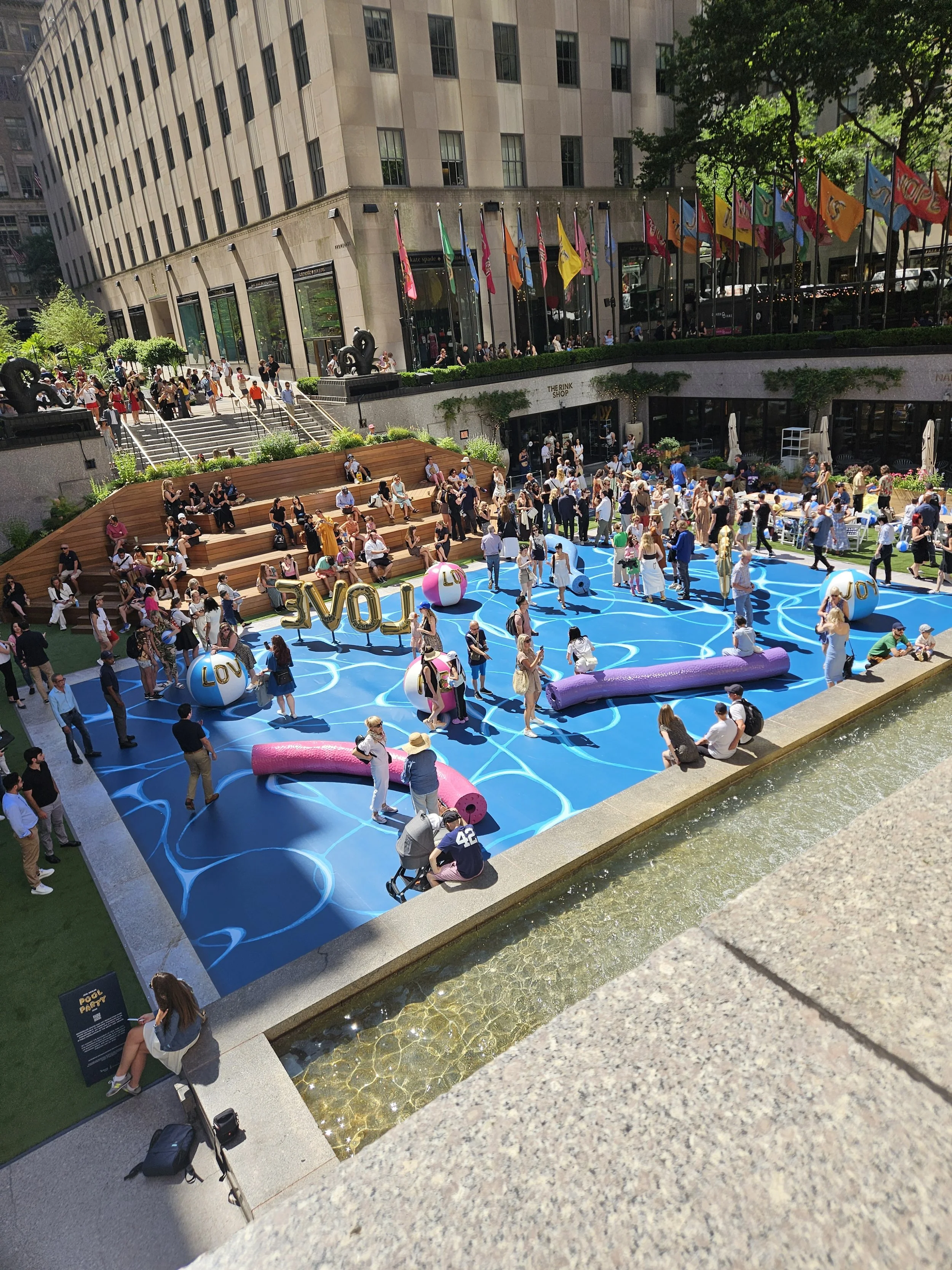 People gathered around a vibrant outdoor pool area on a city street with building and flags in the background.