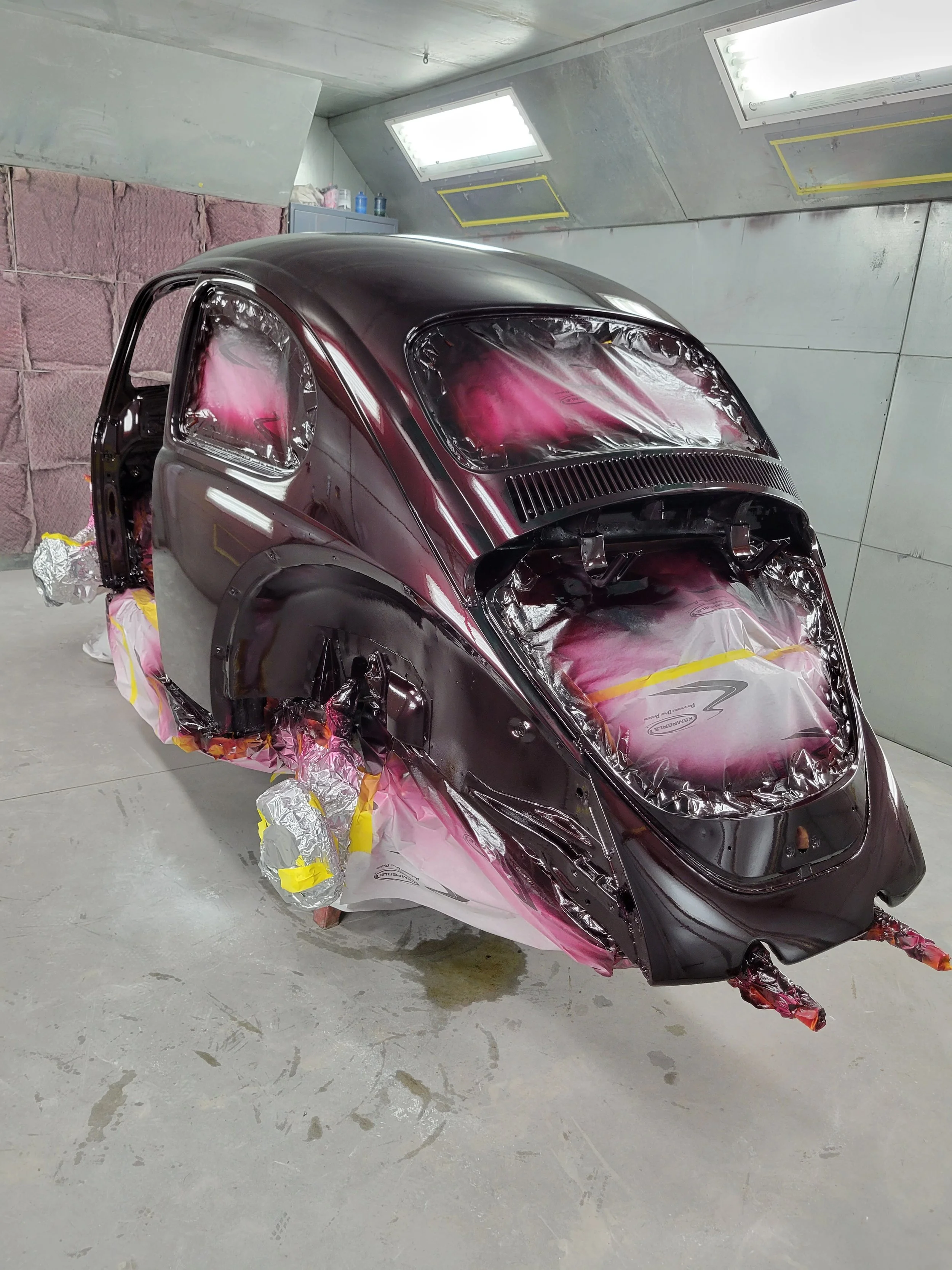 A car body in a paint booth, partially painted dark red, with the windows covered in protective plastic and masking tape. The vehicle is elevated on supports for painting.