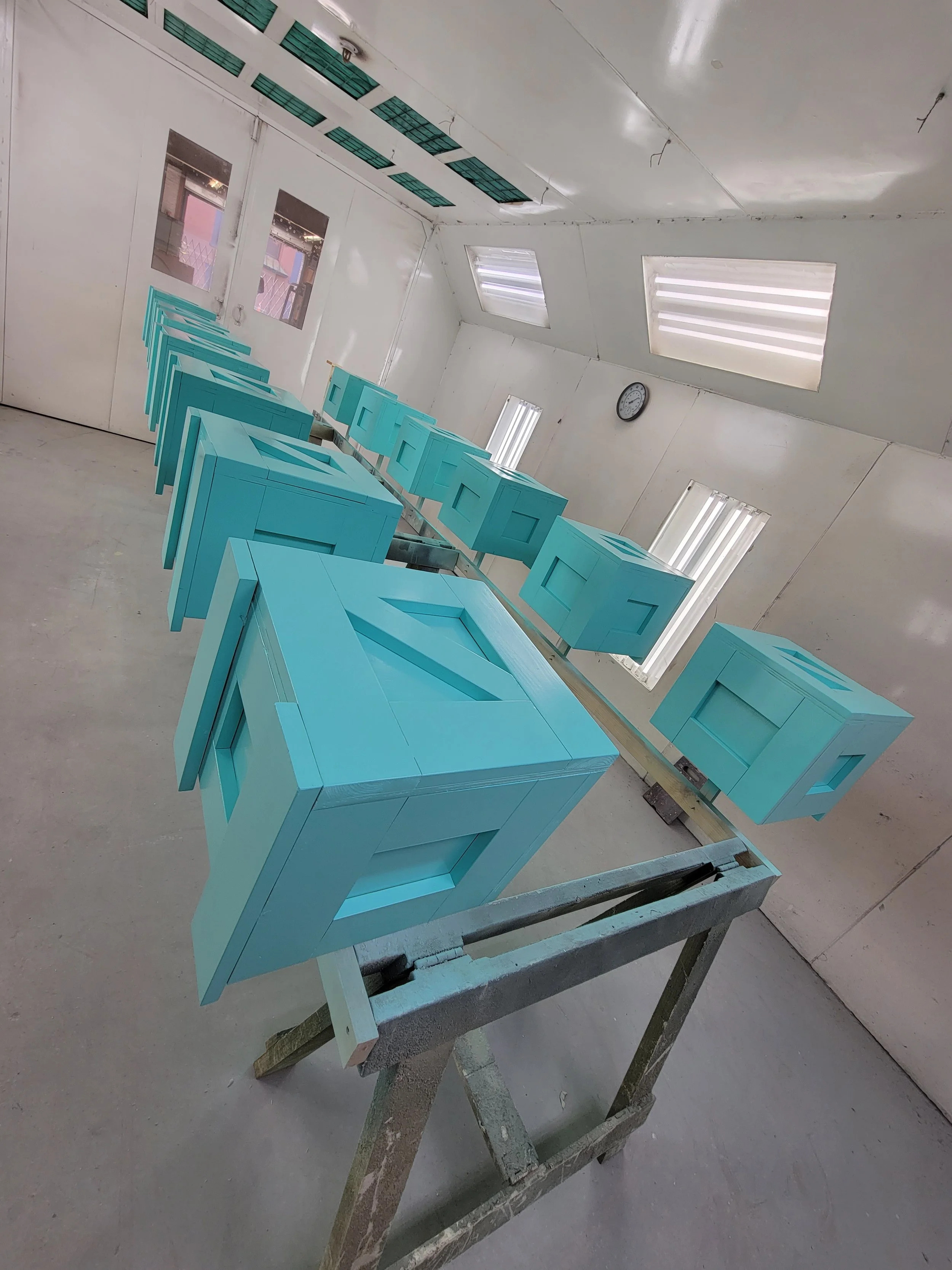 Multiple blue wooden ballot boxes are arranged on a workbench inside a white room with fluorescent lights and a clock on the wall.