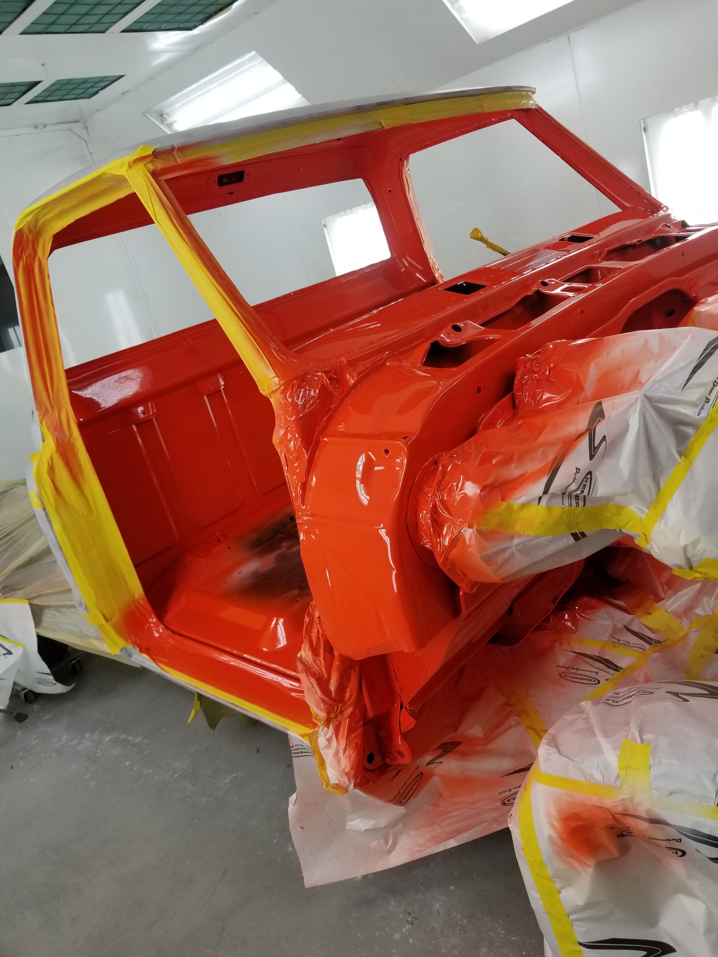 The image shows a vintage vehicle body being painted bright orange in a workshop, with masked areas covered in protective paper and tape.