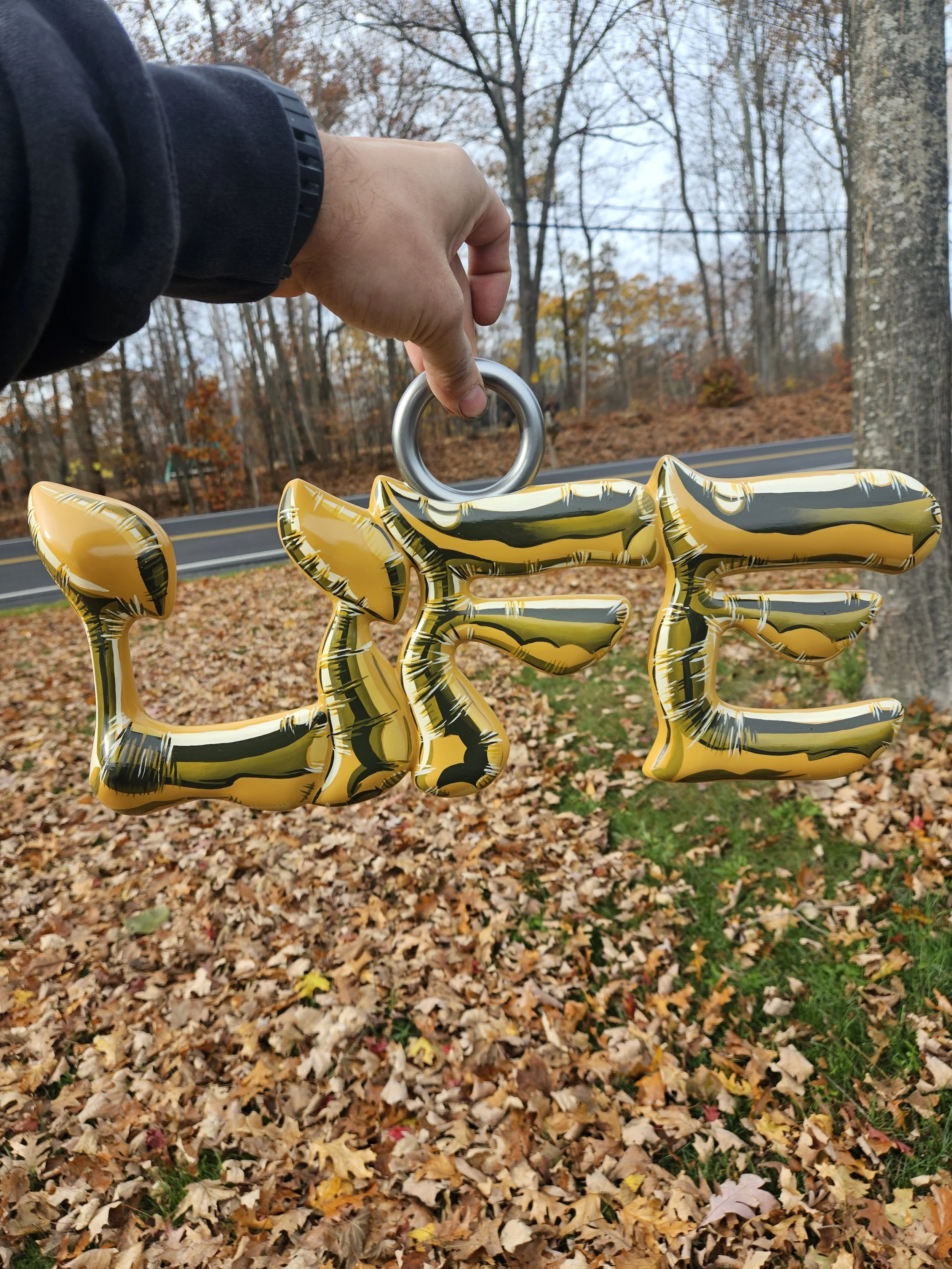 A hand holding a metallic ring attached to a metallic gold and black inflatable decoration spelling out the word "LOVE" against an outdoor background with trees and fallen autumn leaves.