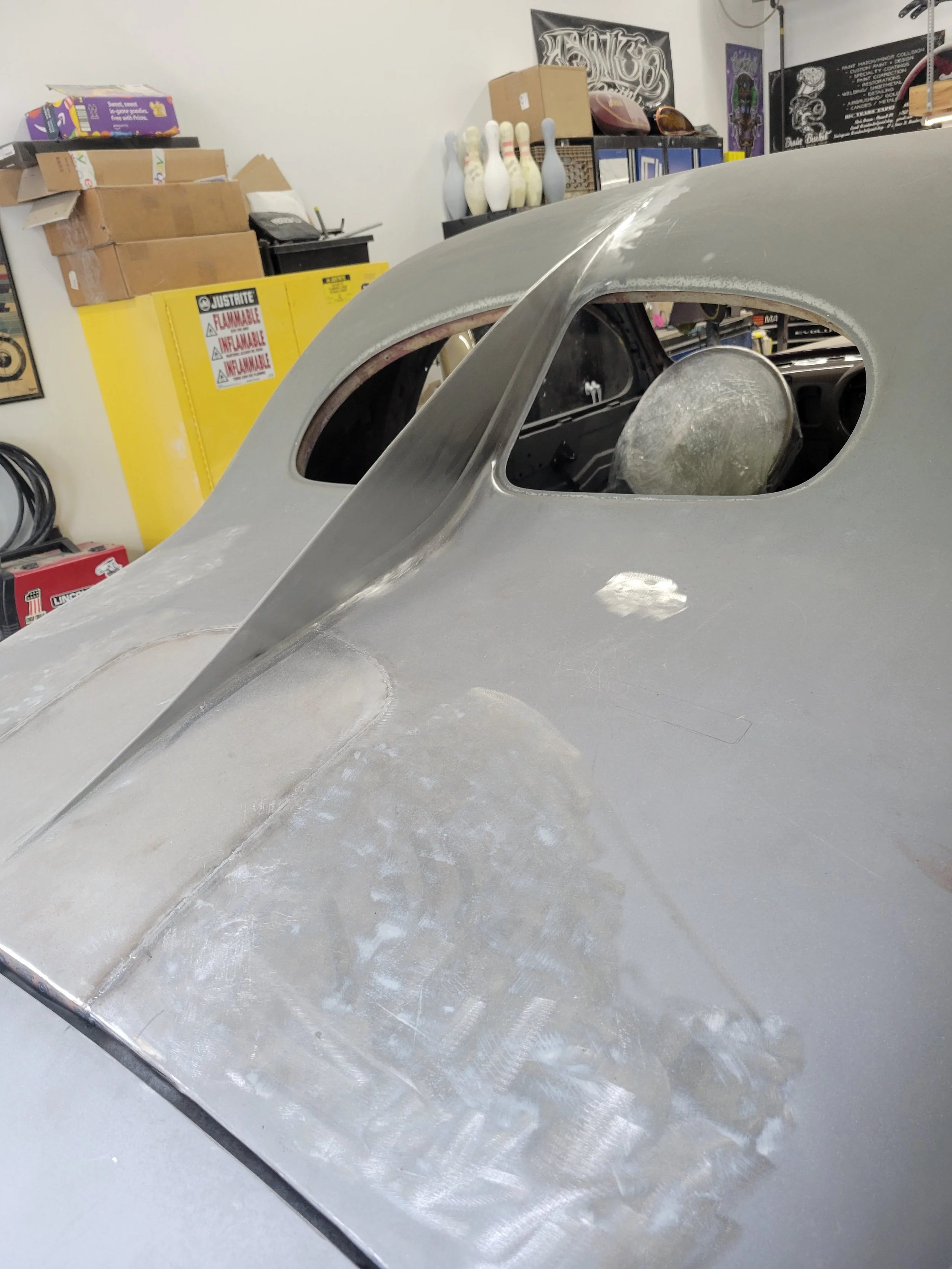 A vintage race car body in the process of restoration, showing an unfinished aluminum exterior with a cockpit and a retro windshield shape, in a garage workshop.