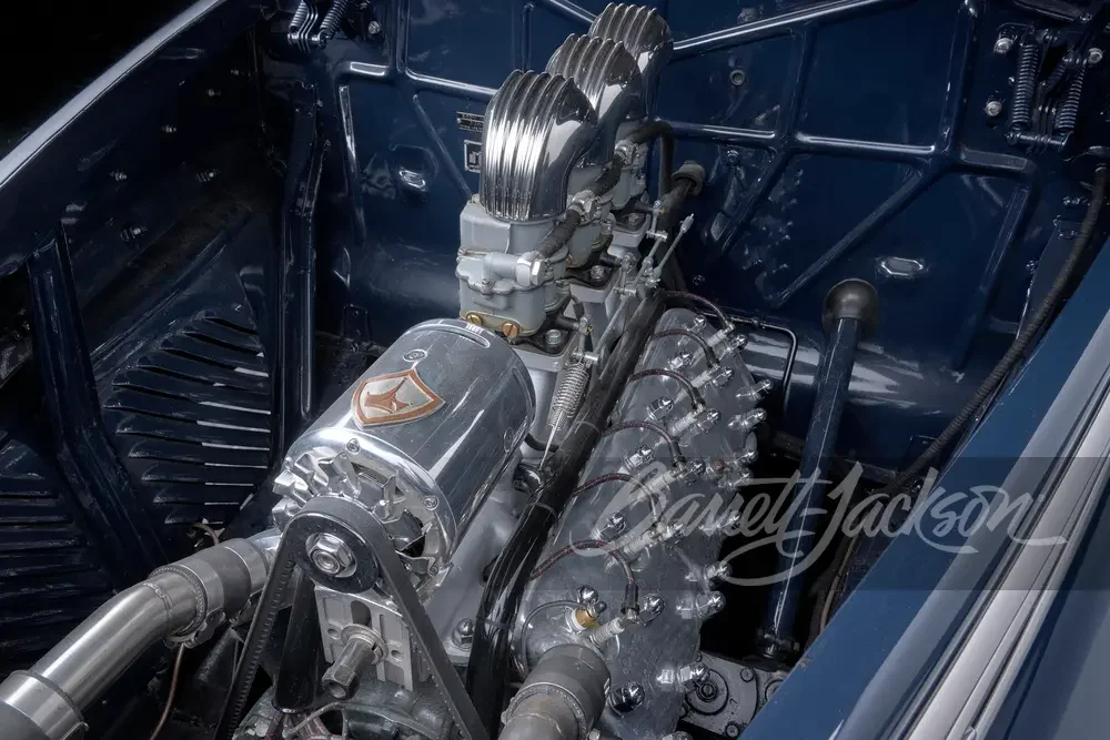 A vintage car engine with polished metal parts, including a supercharger with a badge, and an intake manifold with runners.