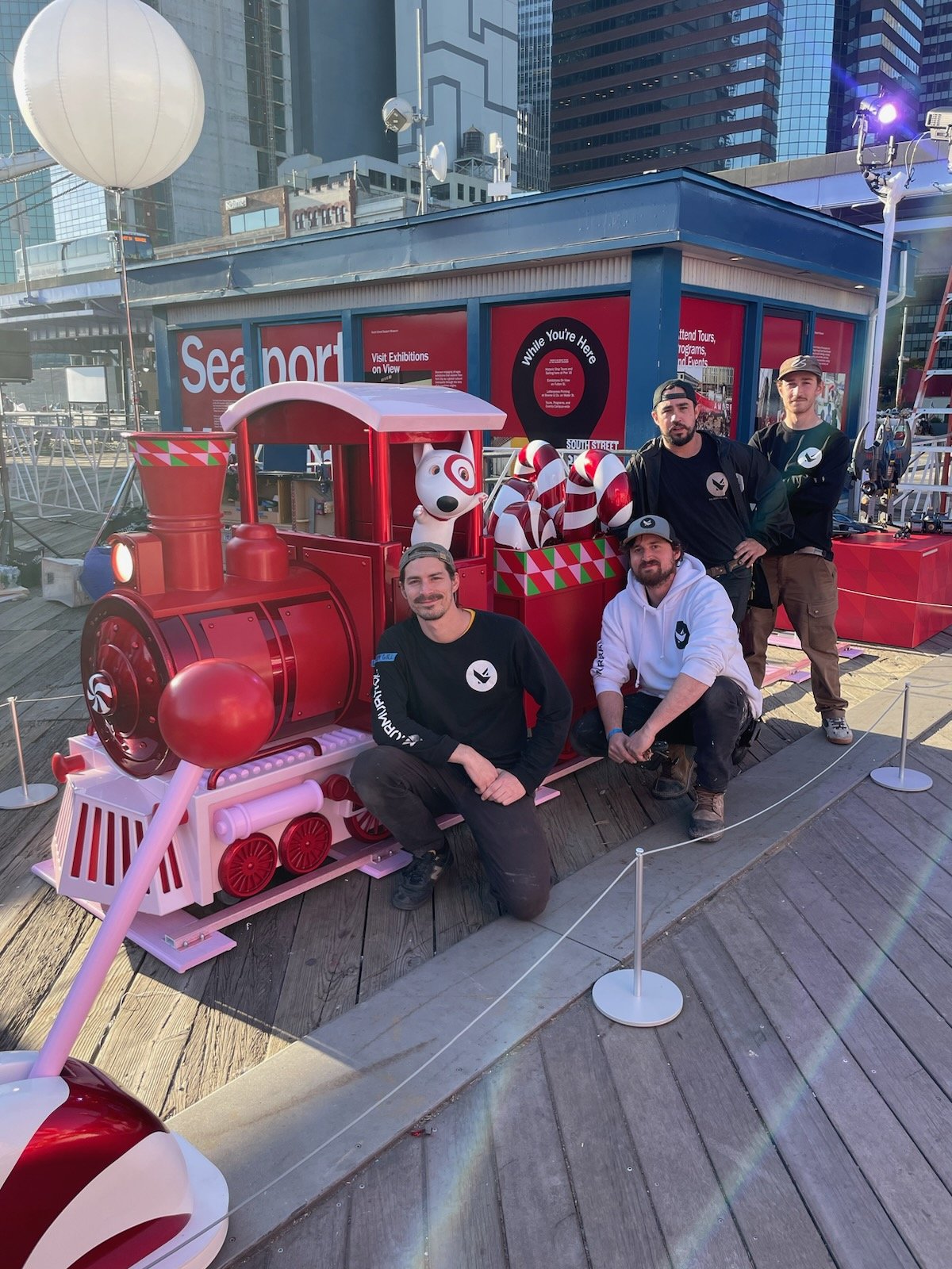 Four men posing in front of a holiday-themed train display on a wooden deck, with city skyscrapers in the background. The train is decorated for Christmas, with red and white candy cane themed elements and a dog character at the front.