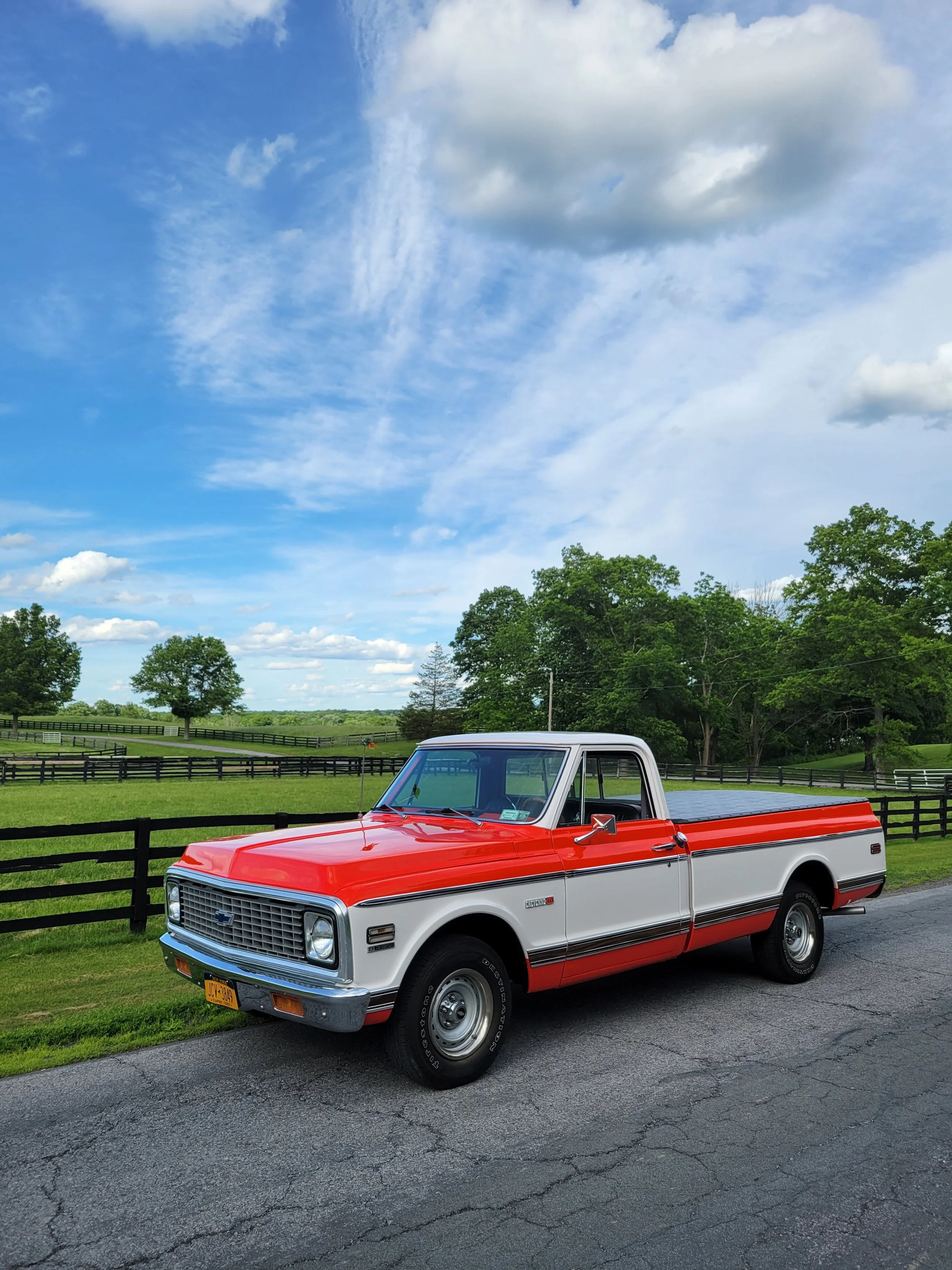 A vintage red and white Chevrolet pickup truck parked on a paved road beside a green field with a wooden fence, under a partly cloudy blue sky.