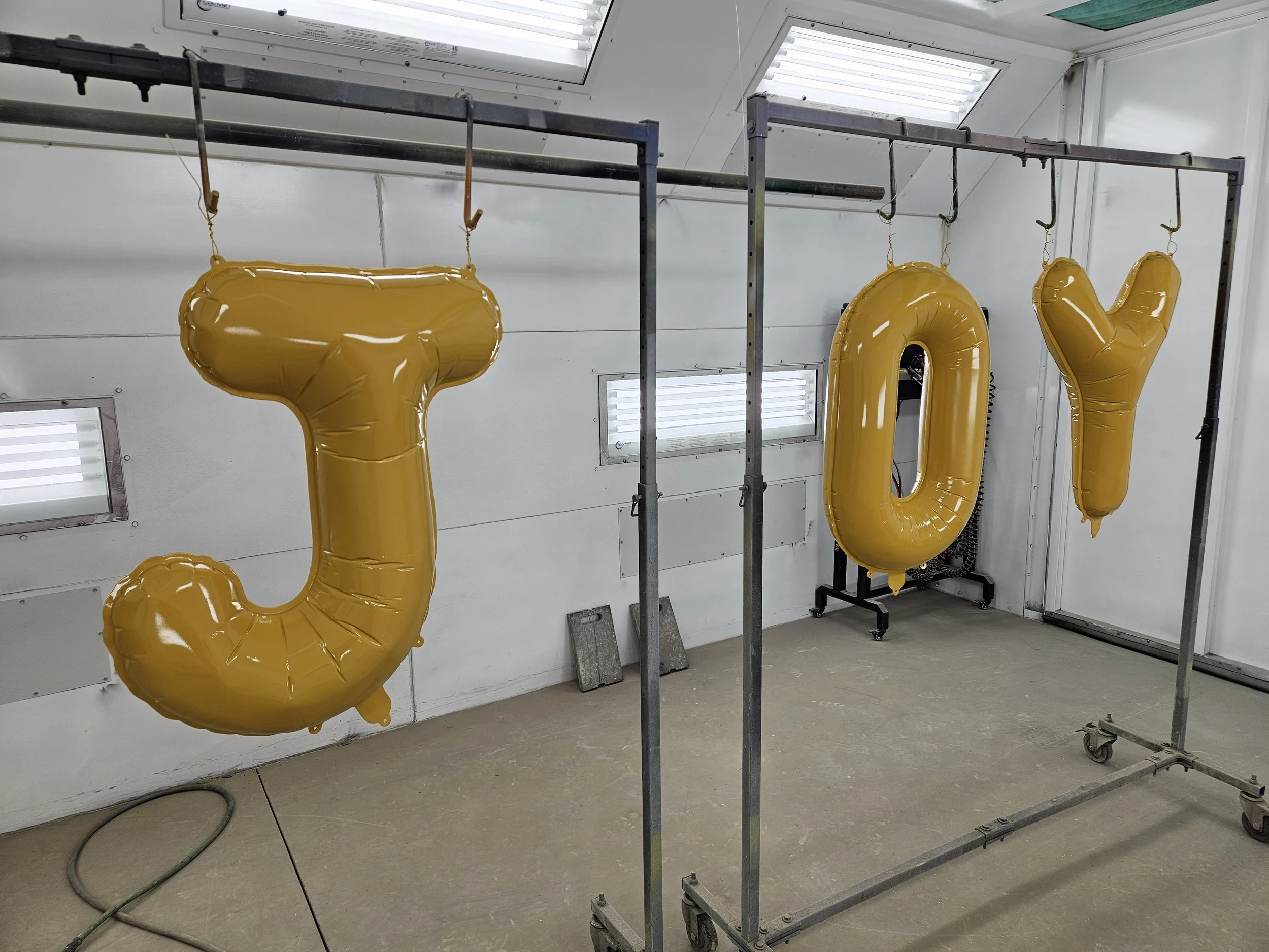 Yellow balloons shaped like the letters J, O, and Y hanging from a metal frame. The setting appears to be a workshop or storage area with white walls and fluorescent lighting.