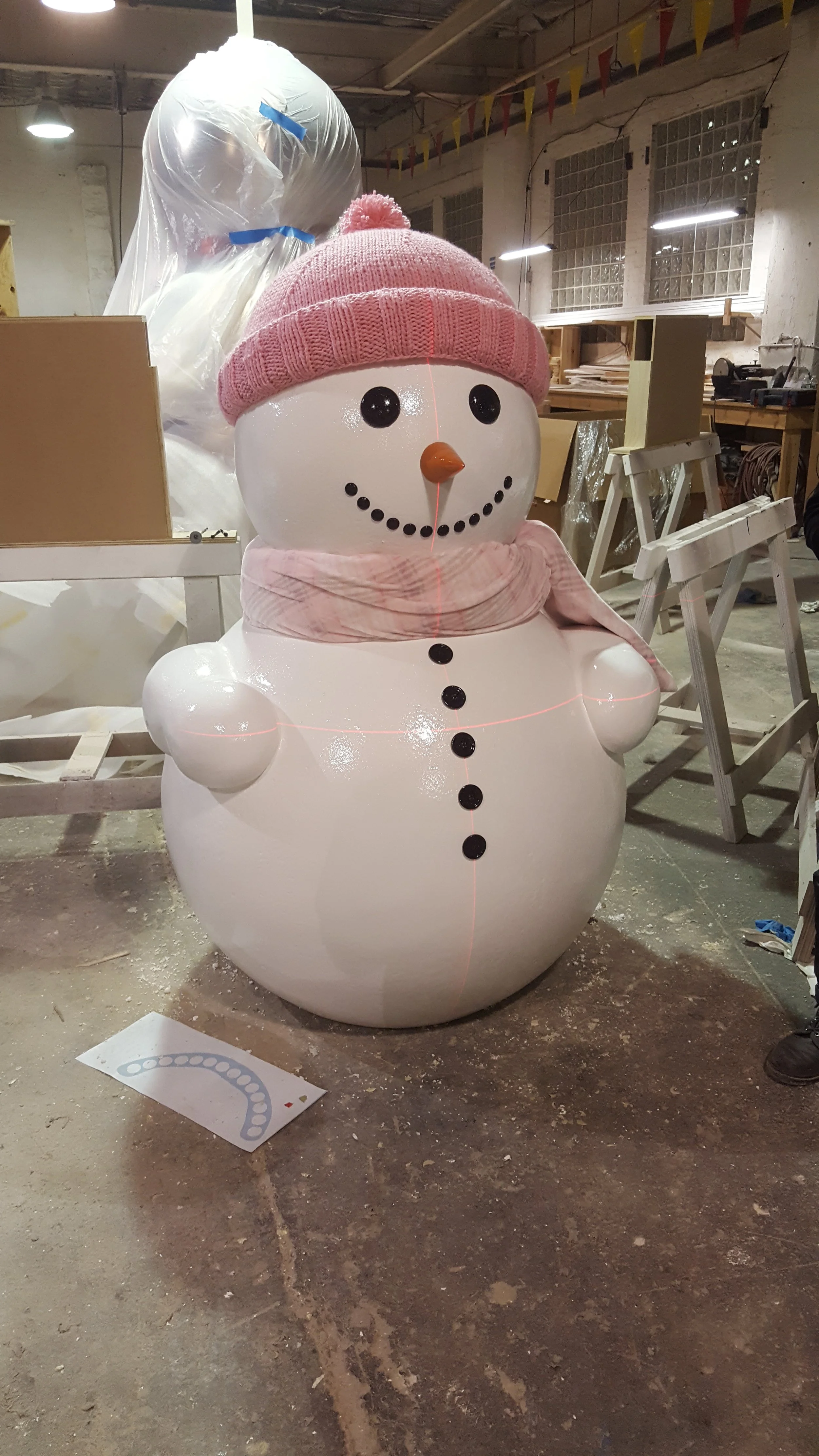 A large snowman decoration with a pink knit hat, matching scarf, black buttons, and a carrot nose, standing in a workshop with various tools and materials around.