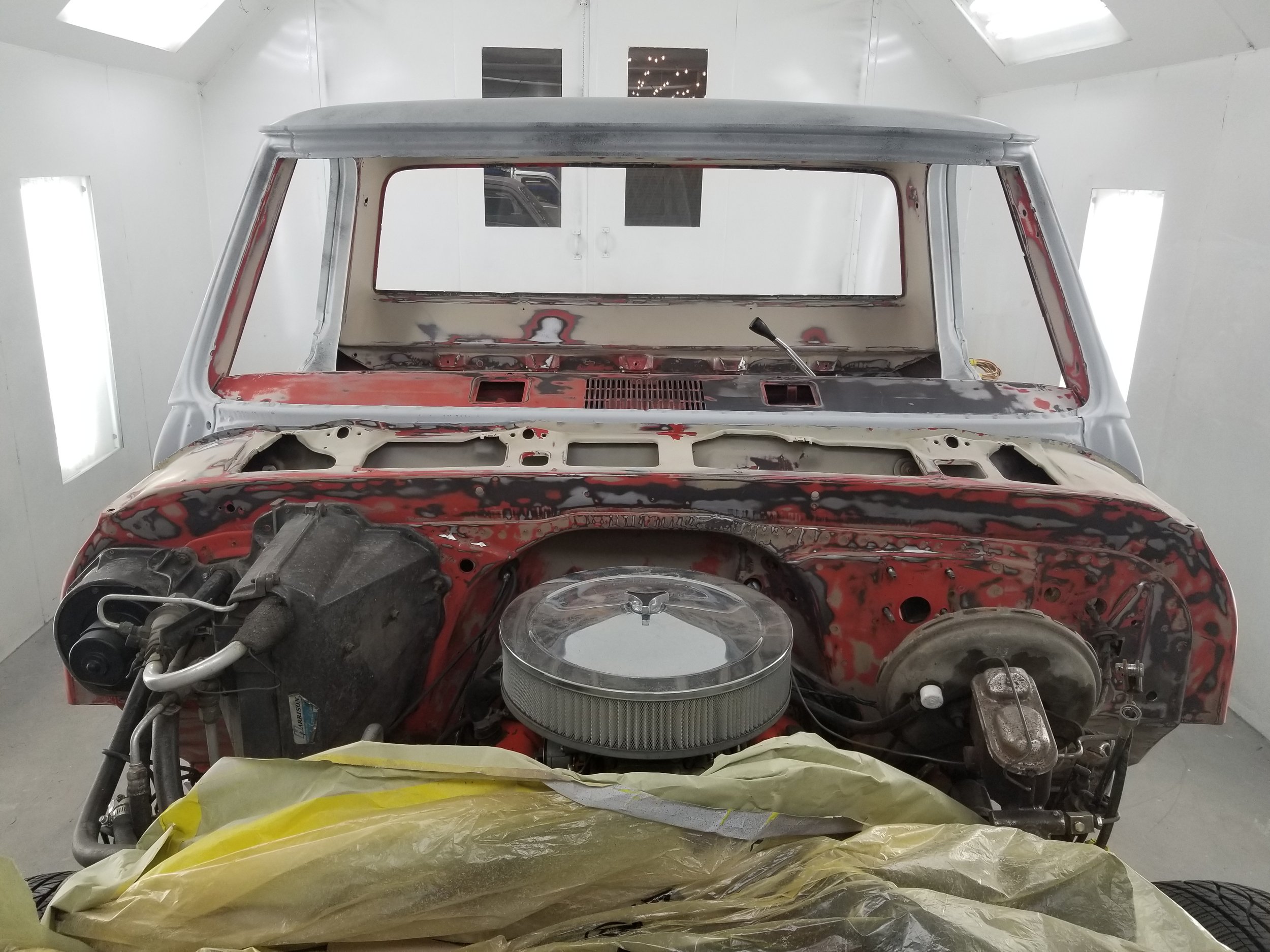 Front view of a vintage car body undergoing restoration inside a white paint booth, with parts of the hood and engine area exposed and unpainted, featuring a large round air filter on the engine and various mechanical components.