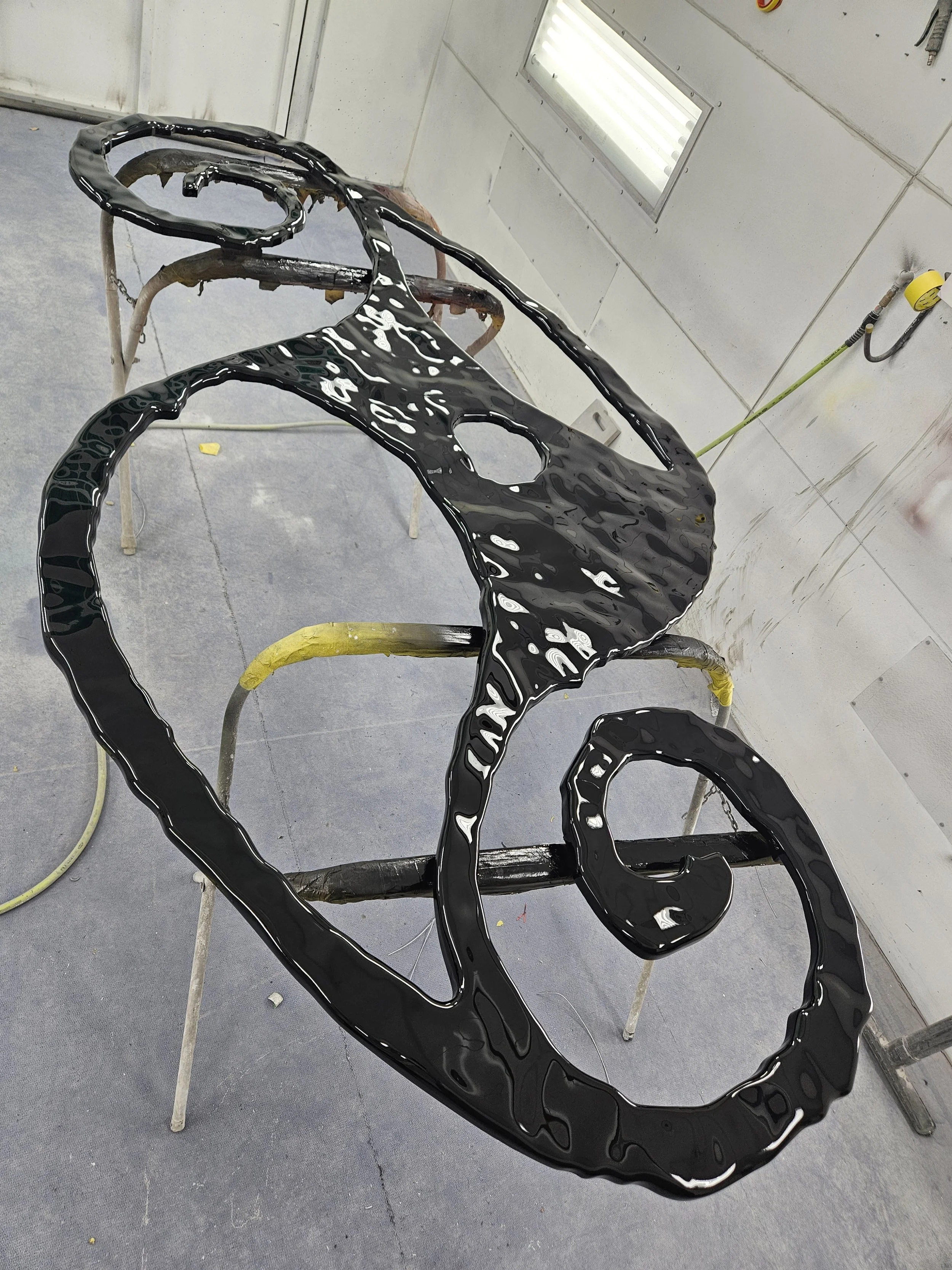 A black metal sculpture of a bicycle on a stand in a workshop, with a window and white walls in the background.