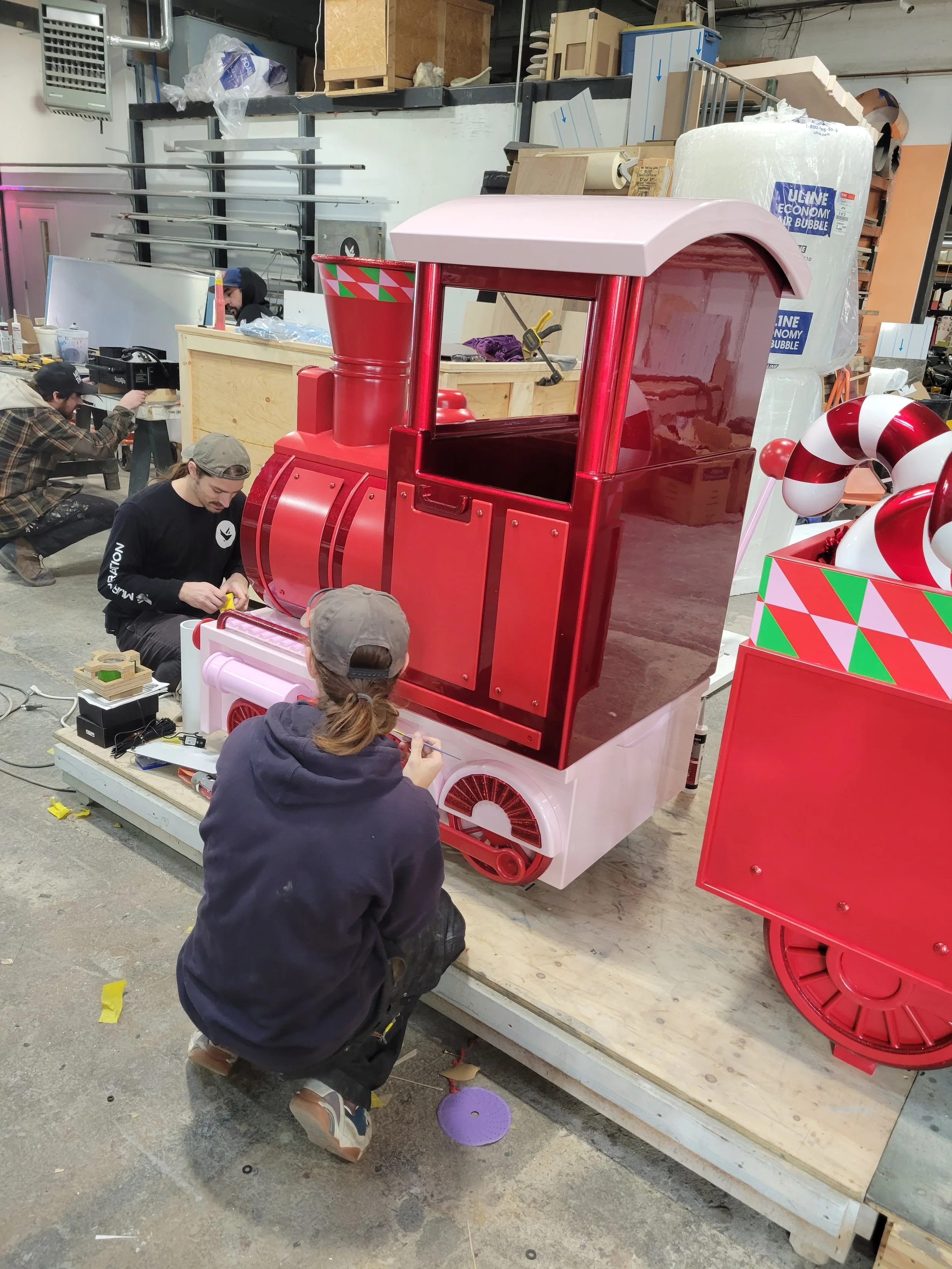 Workers assembling a festive red and pink train-shaped holiday display with candy cane decorations.