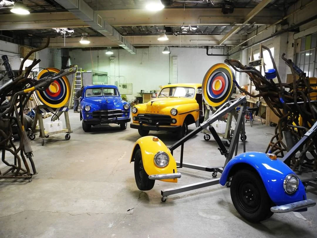 A workshop with three vintage cars, two are yellow and one is blue, and various bicycle frames and wheel components on stands and racks.