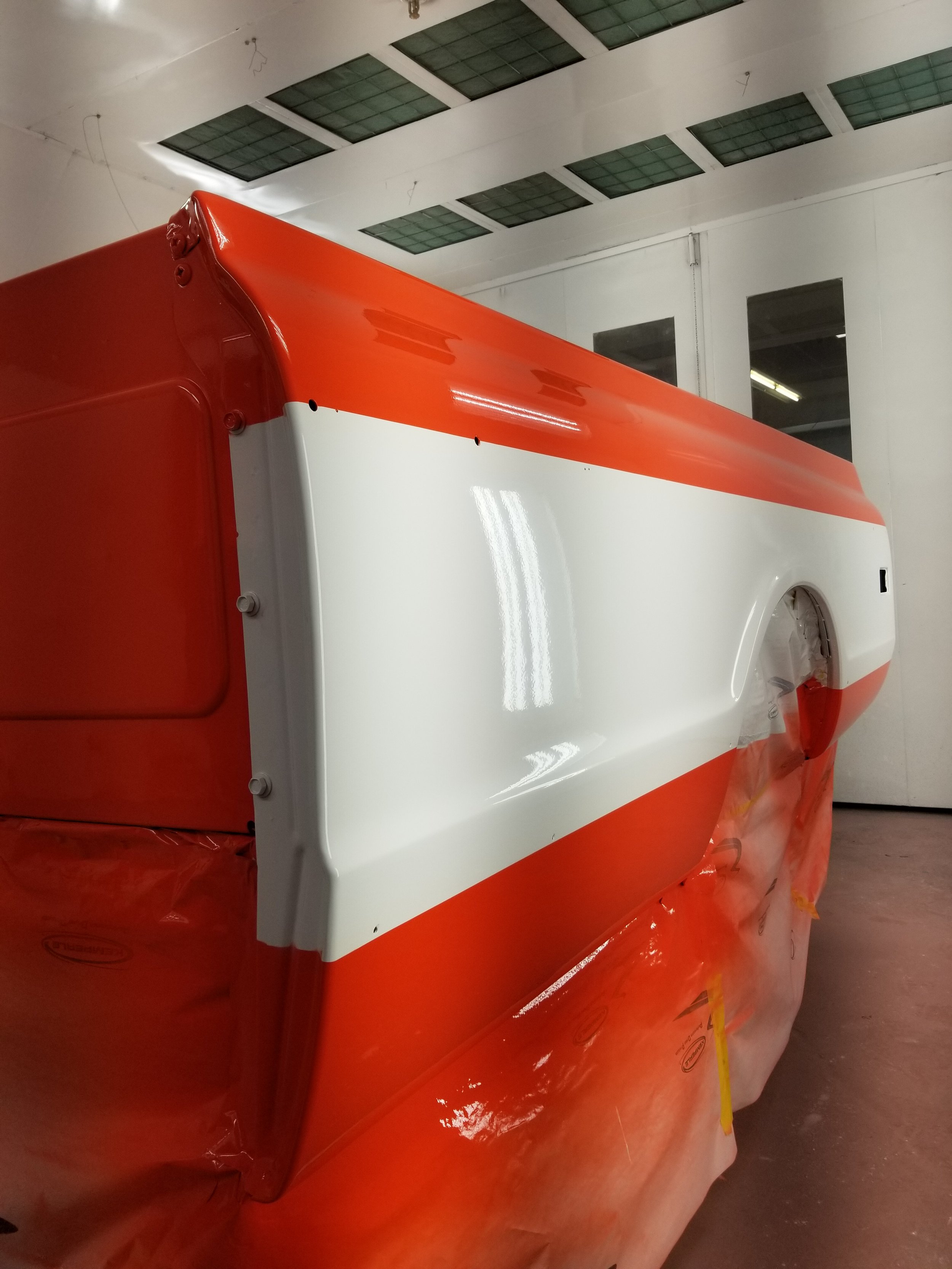 A vehicle's body painted in orange and white in a workshop, with some parts covered and protected with plastic for painting.