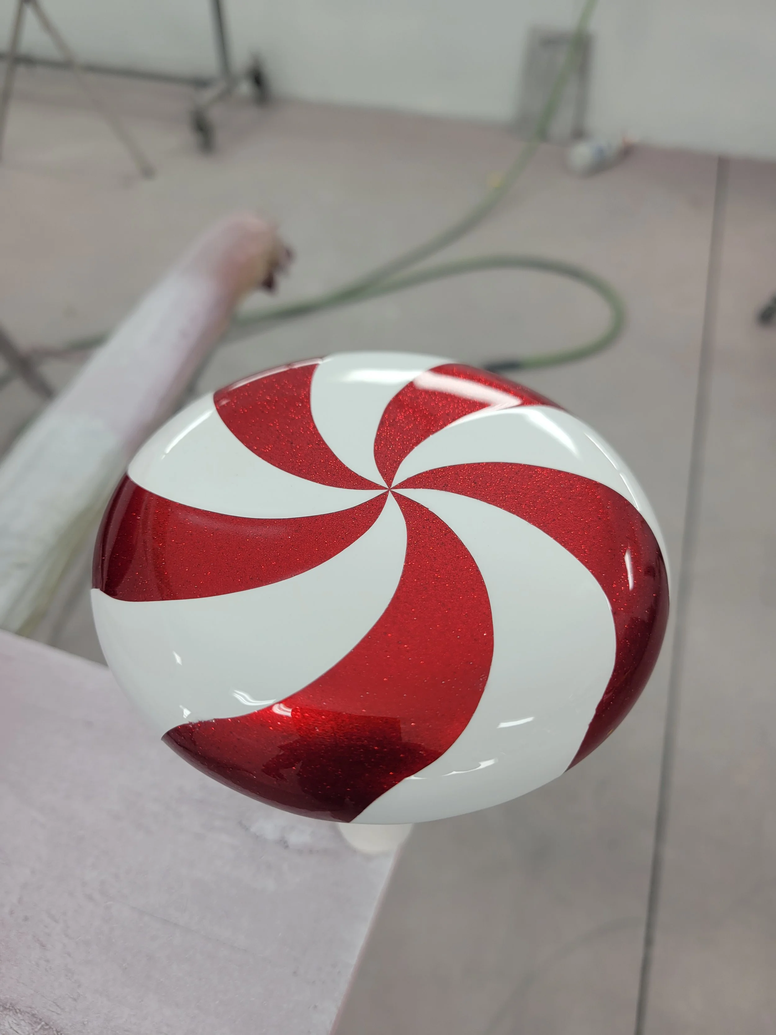A spherical peppermint-themed ornament with red and white swirls and a glittery finish, held above a light-colored surface in an indoor setting.