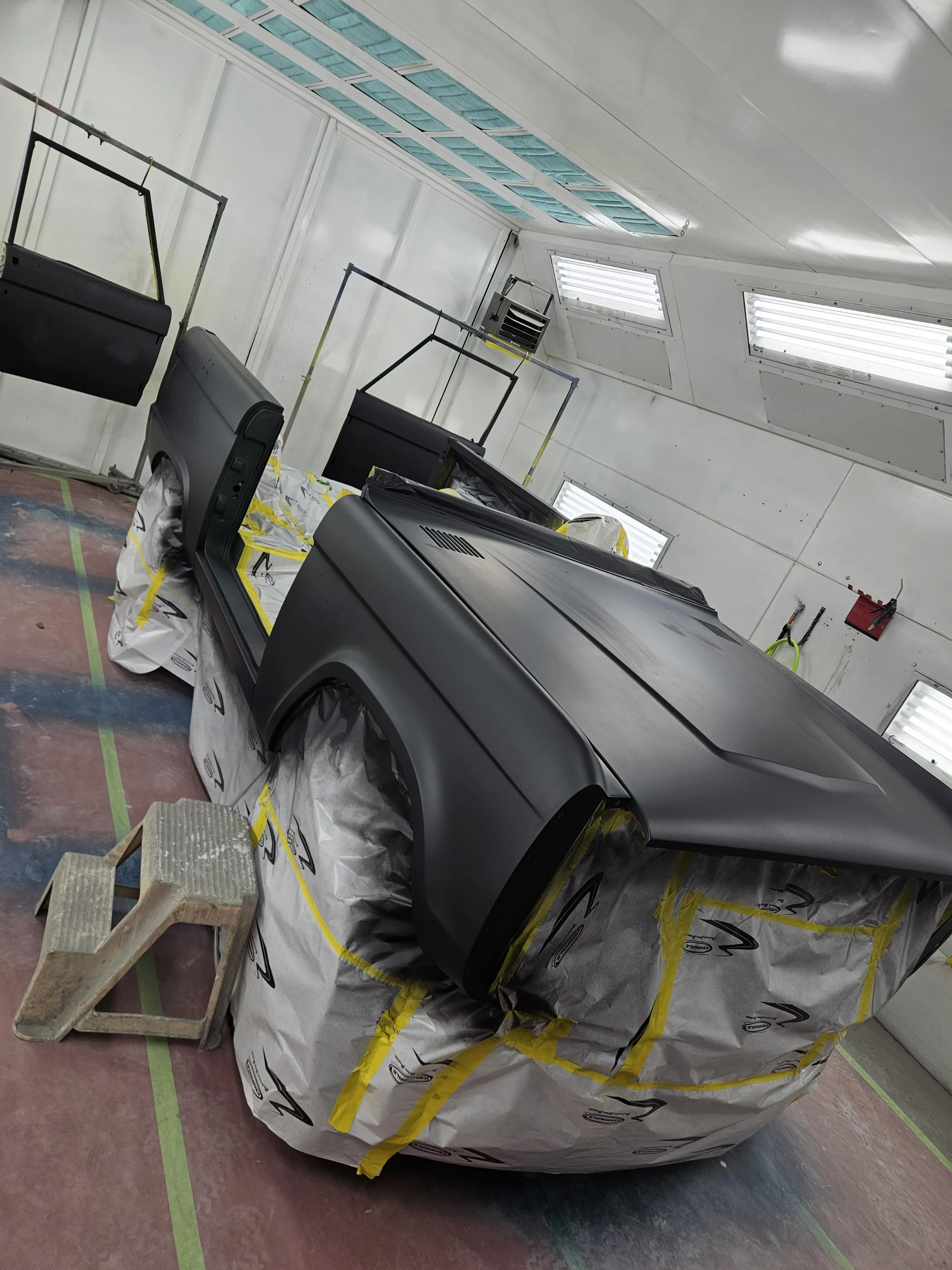 A vehicle in a paint booth, with its body painted matte black, partially masked with paper and tape, lying on its side, with some masking paper on the floor and a small step stool nearby.