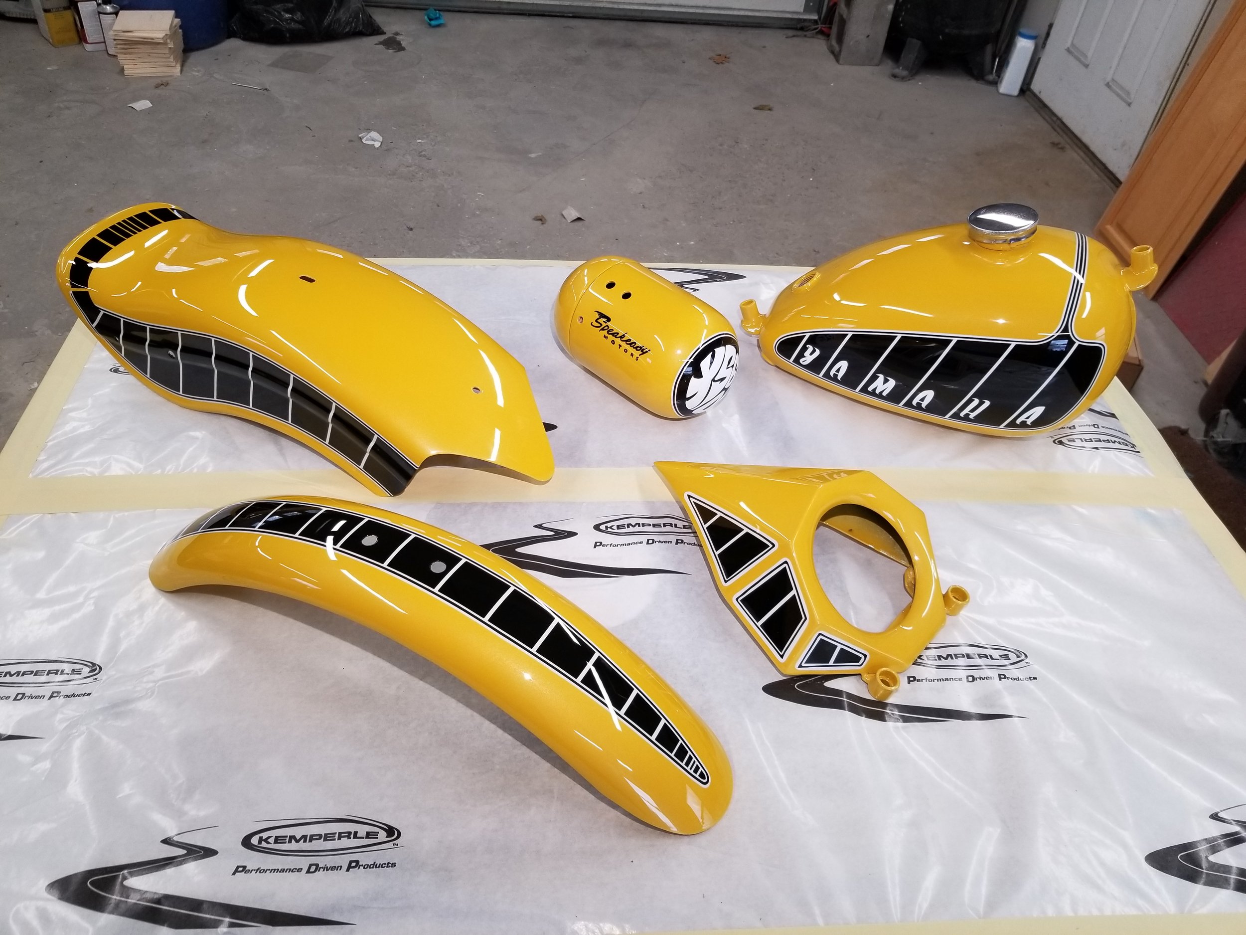 Yellow motorcycle fuel tank, seat cowl, and other motorcycle parts laid on a white surface with Kelperle branding, in a garage setting.