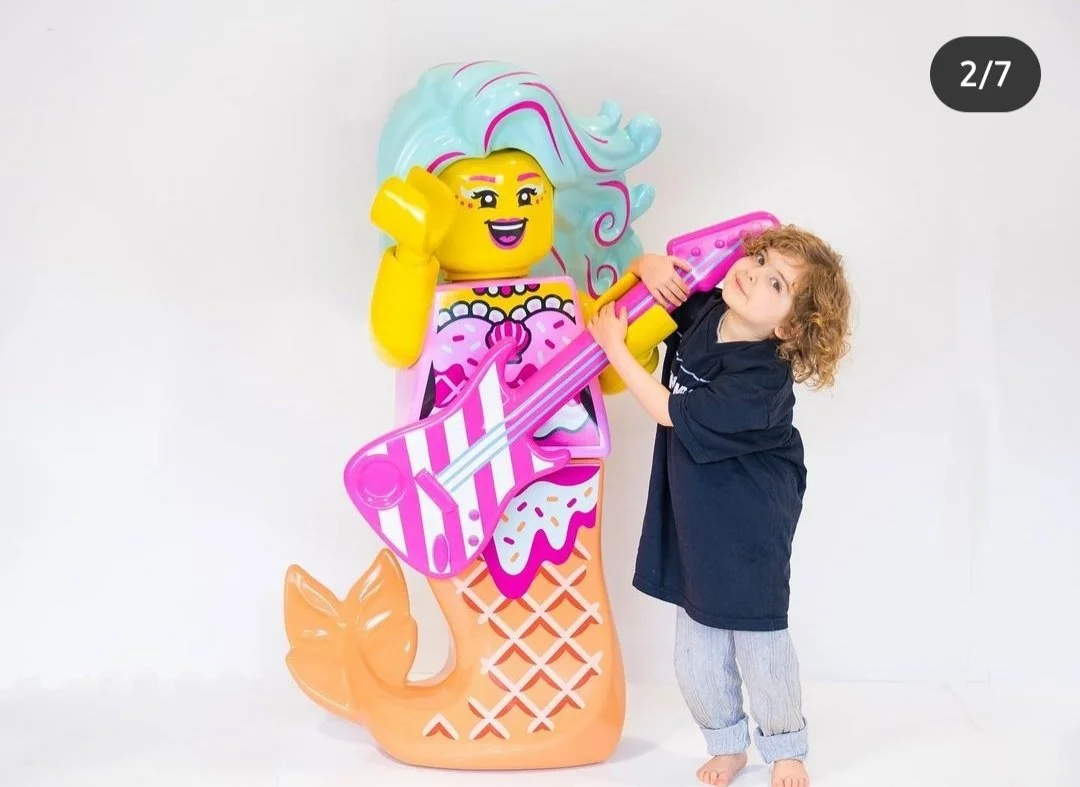 A young child with curly hair holding a yellow bubble wand poses next to a large LEGO mermaid with blue hair, pink fins, and a tail, in front of a plain white background.