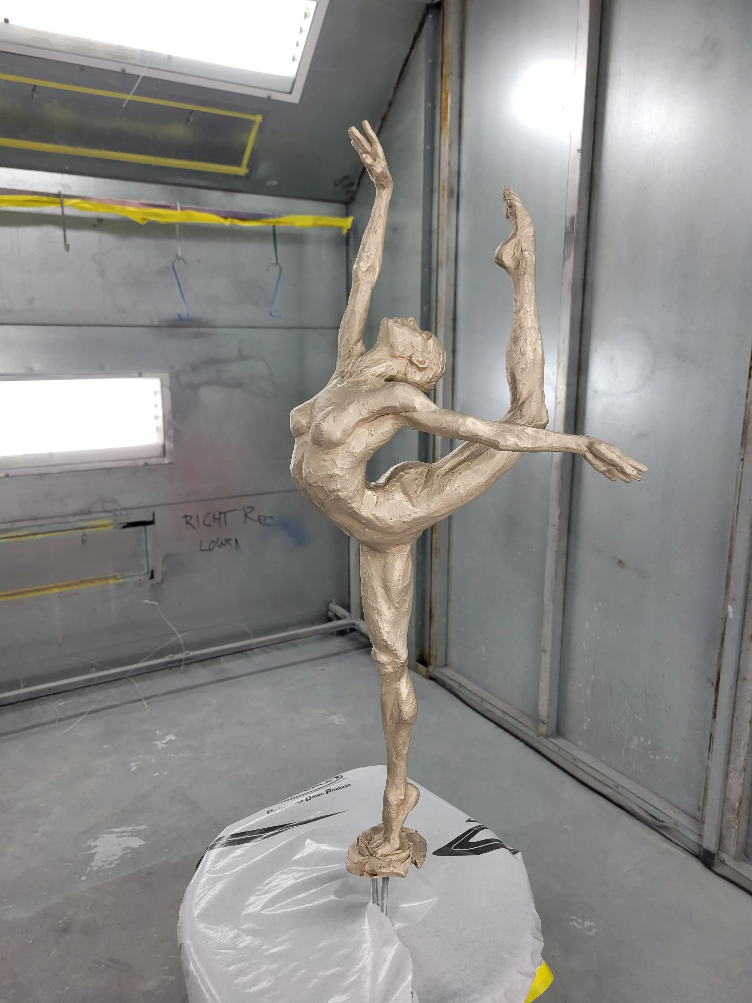 A metallic sculpture of a dancer in an elegant pose with one leg lifted and arms extended, set in an industrial setting with metal walls and a window.