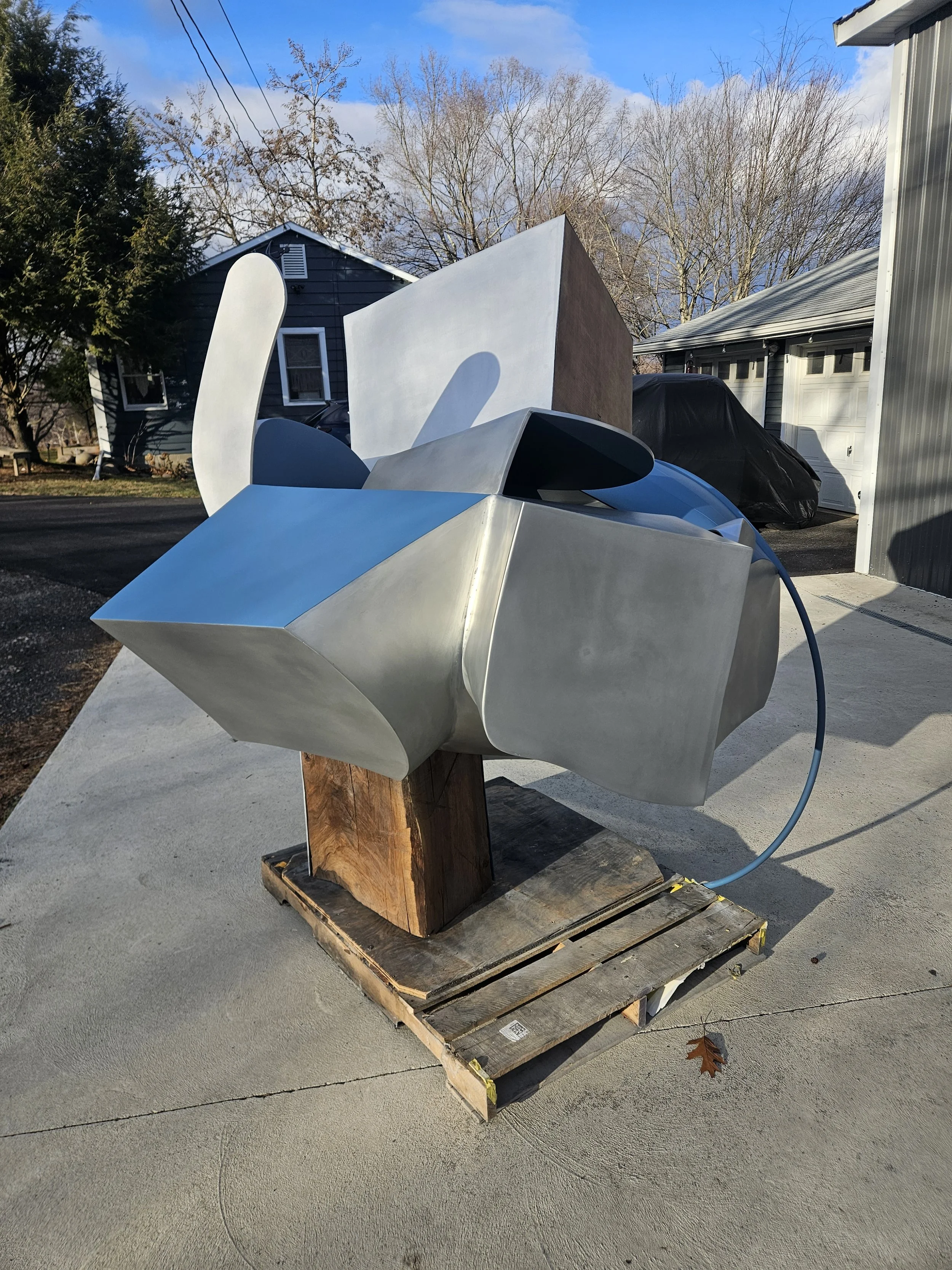 Abstract metal sculpture mounted on a wooden base outdoors, with a house and trees in the background.