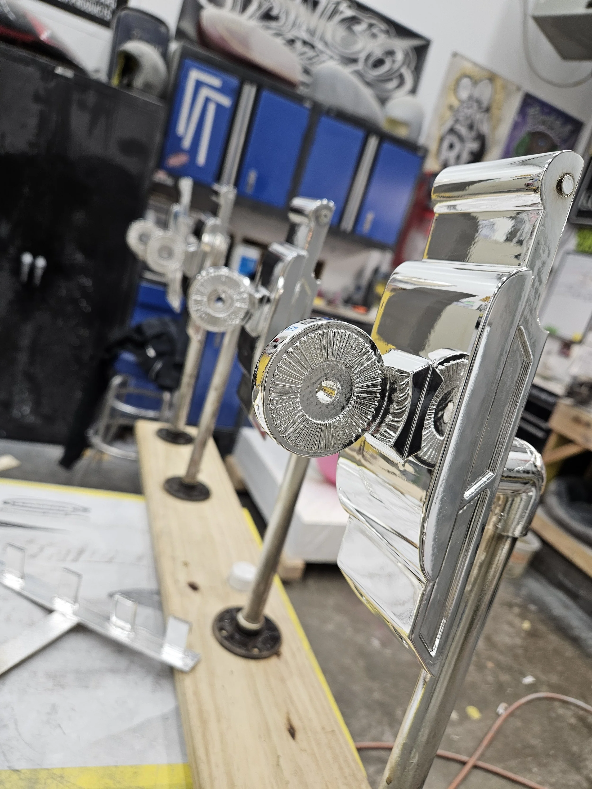 Close-up of a metal drill press in a workshop, with a wooden base support, surrounded by tools and equipment in the background.