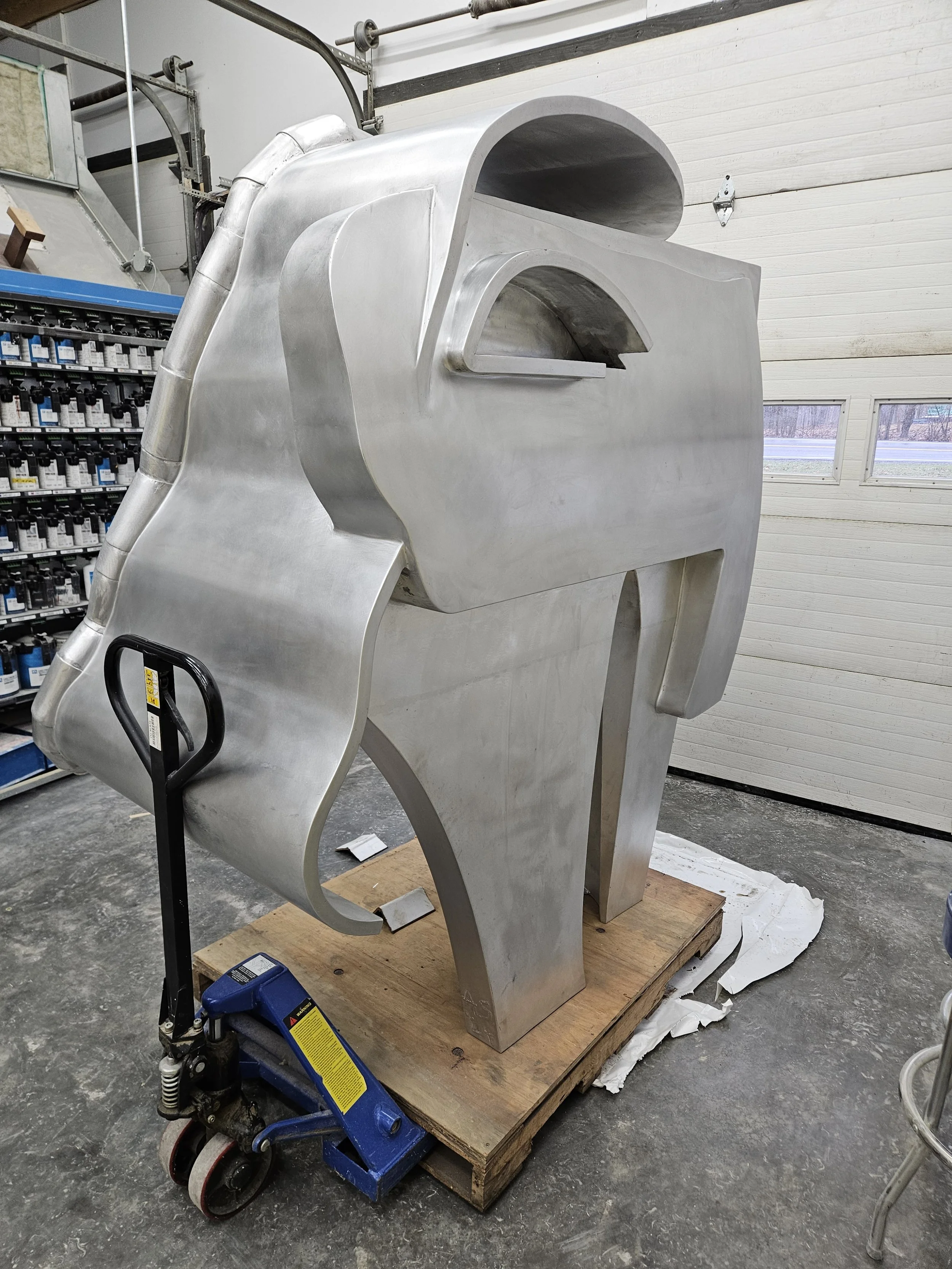 A large, unfinished metallic sculpture resembling a robot or character with a boxy head and body, standing on a wooden pallet in a workshop.