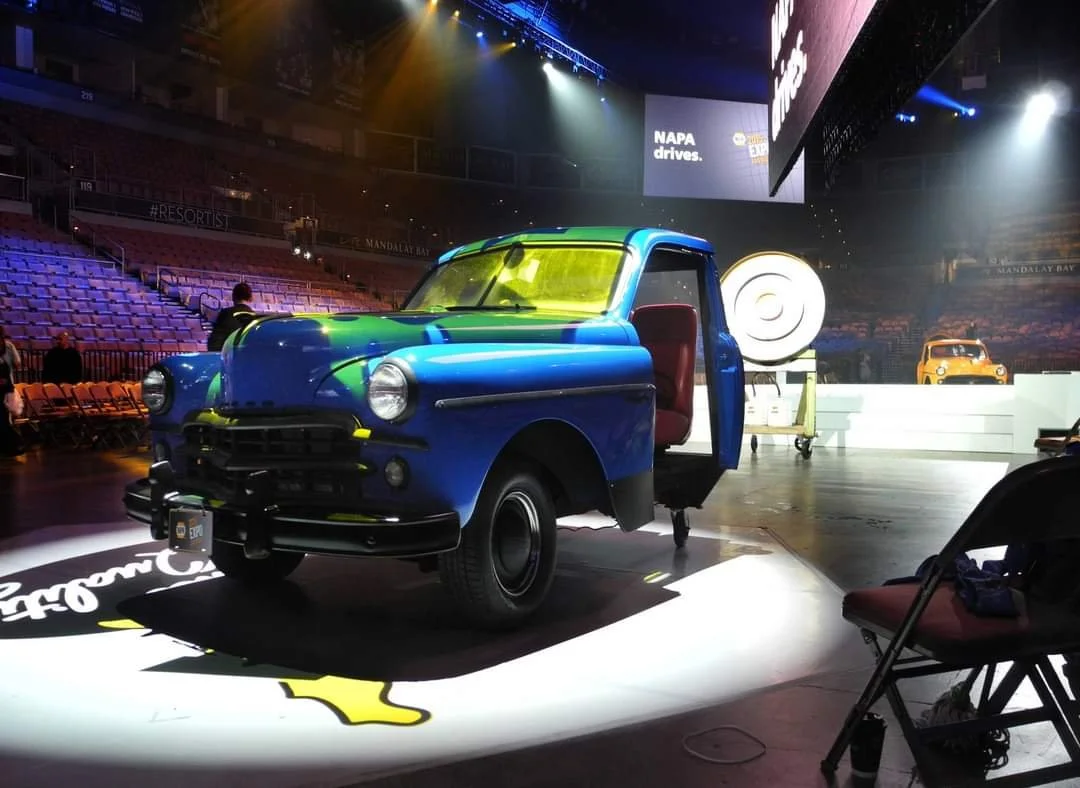 A vintage blue and green van with the driver's side door open, displayed on a stage at an event with bright lights, around rows of empty chairs and a large screen with the text "NAPA drives".
