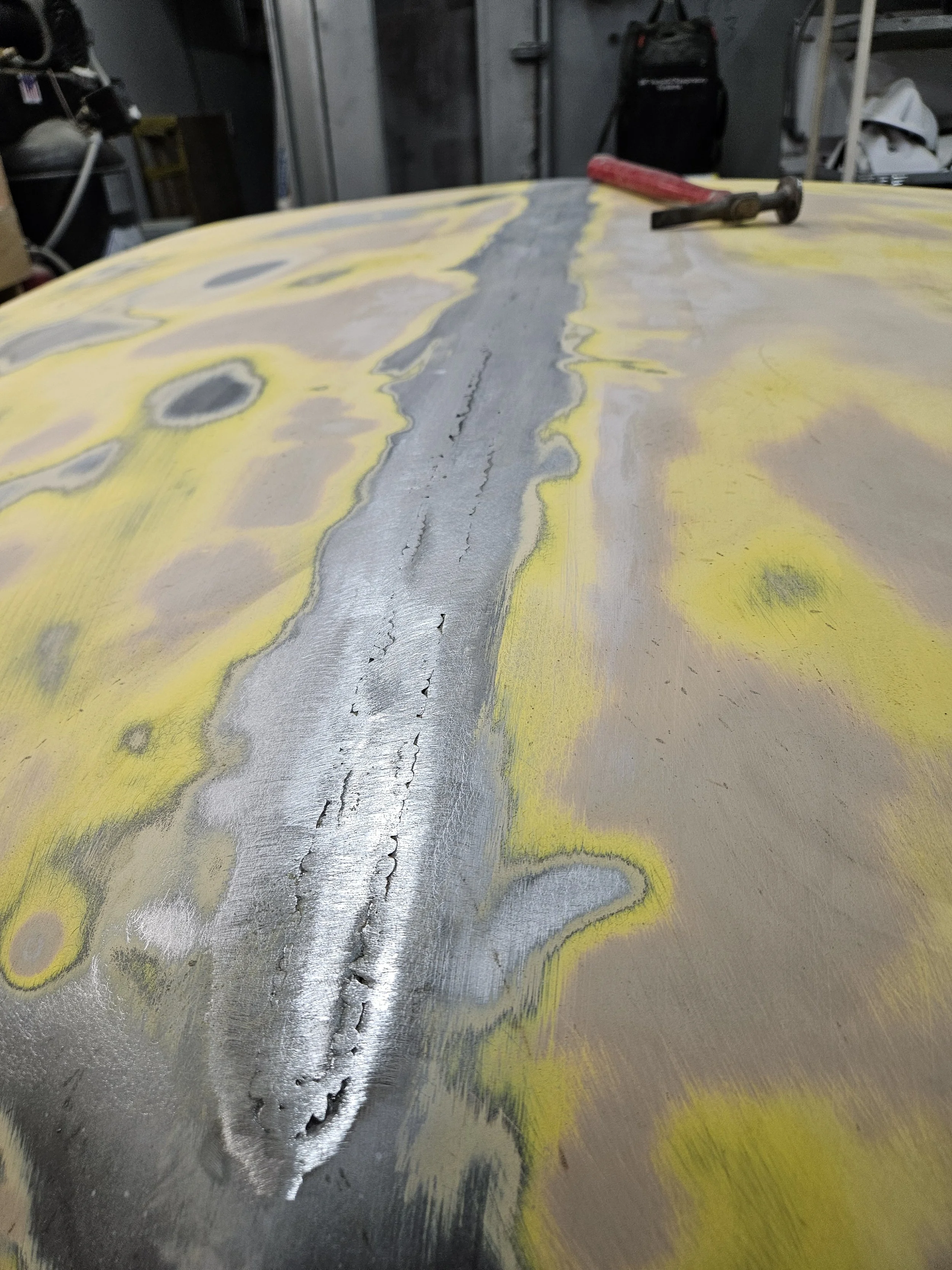 Close-up of a vehicle's surface under repair, showing a large crack filled with seam sealer, with yellow and brown sanding marks around it, and tools like a hammer and impact driver on the surface.