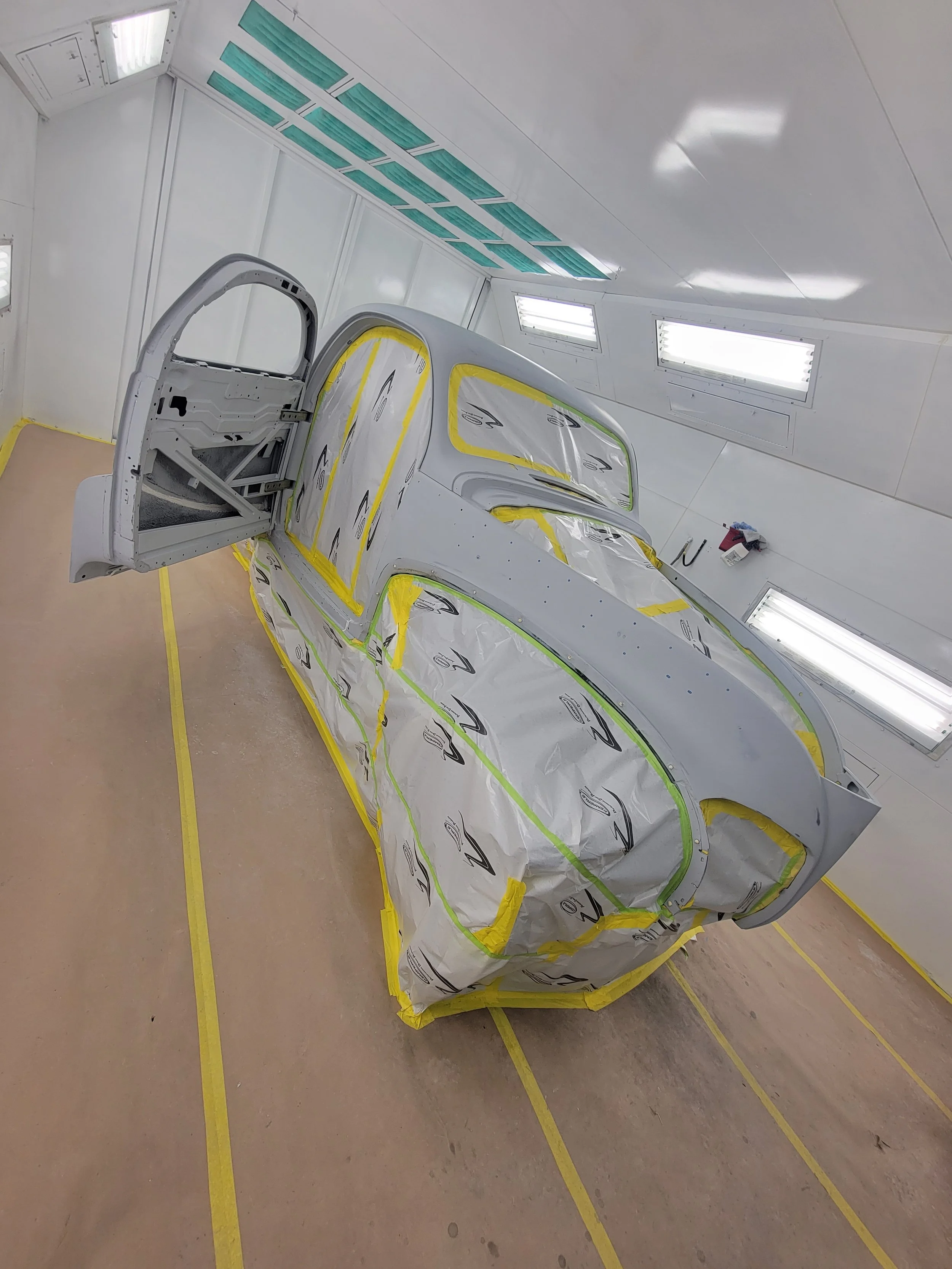 A car body in a paint booth, partially covered with protective plastic and masking tape, with the doors and windows removed. The booth is well-lit with fluorescent lights.