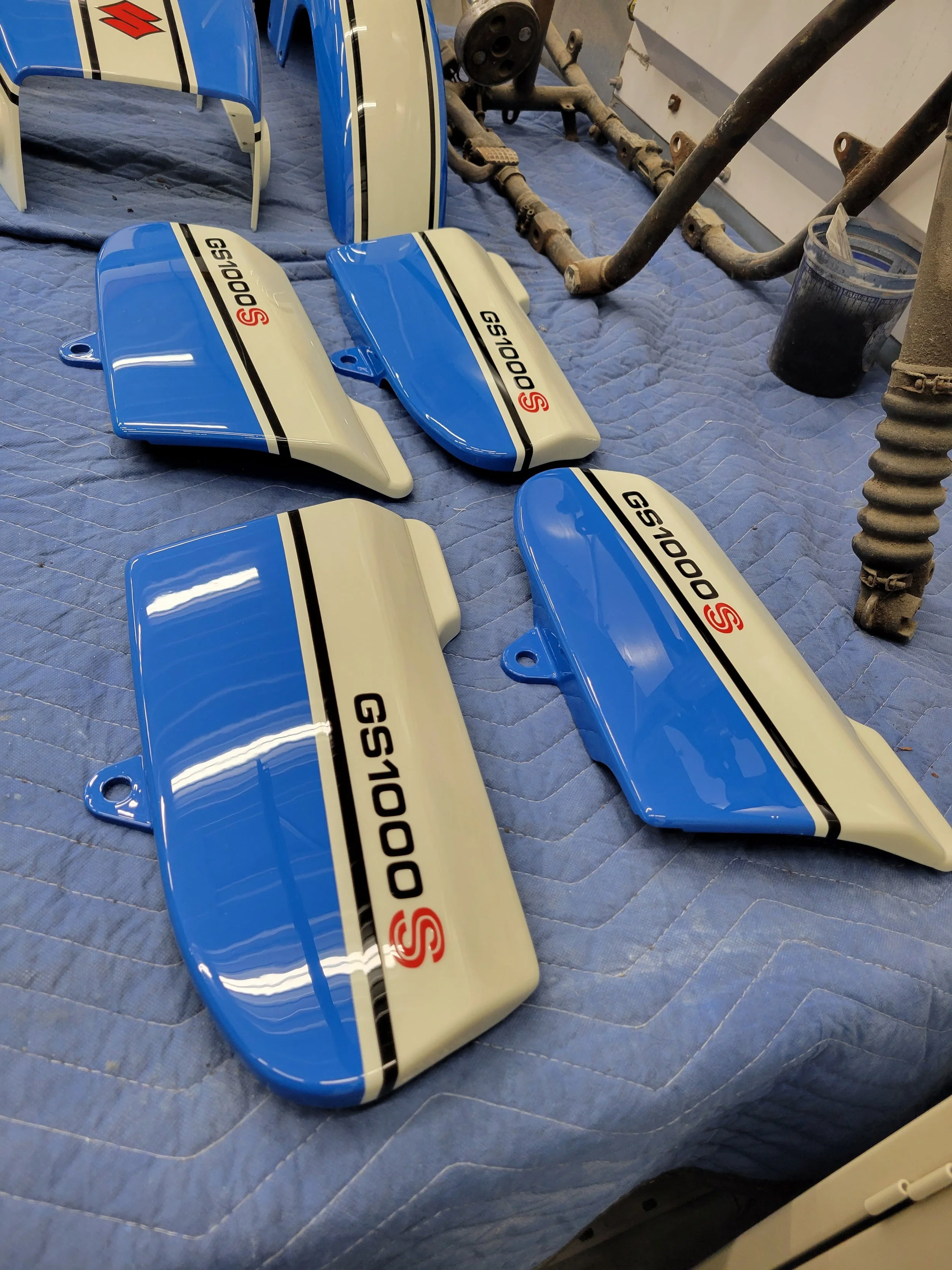 Four blue and white motorcycle tank panels with 'GS1000S' logo laid out on a blue quilted surface in a workshop.