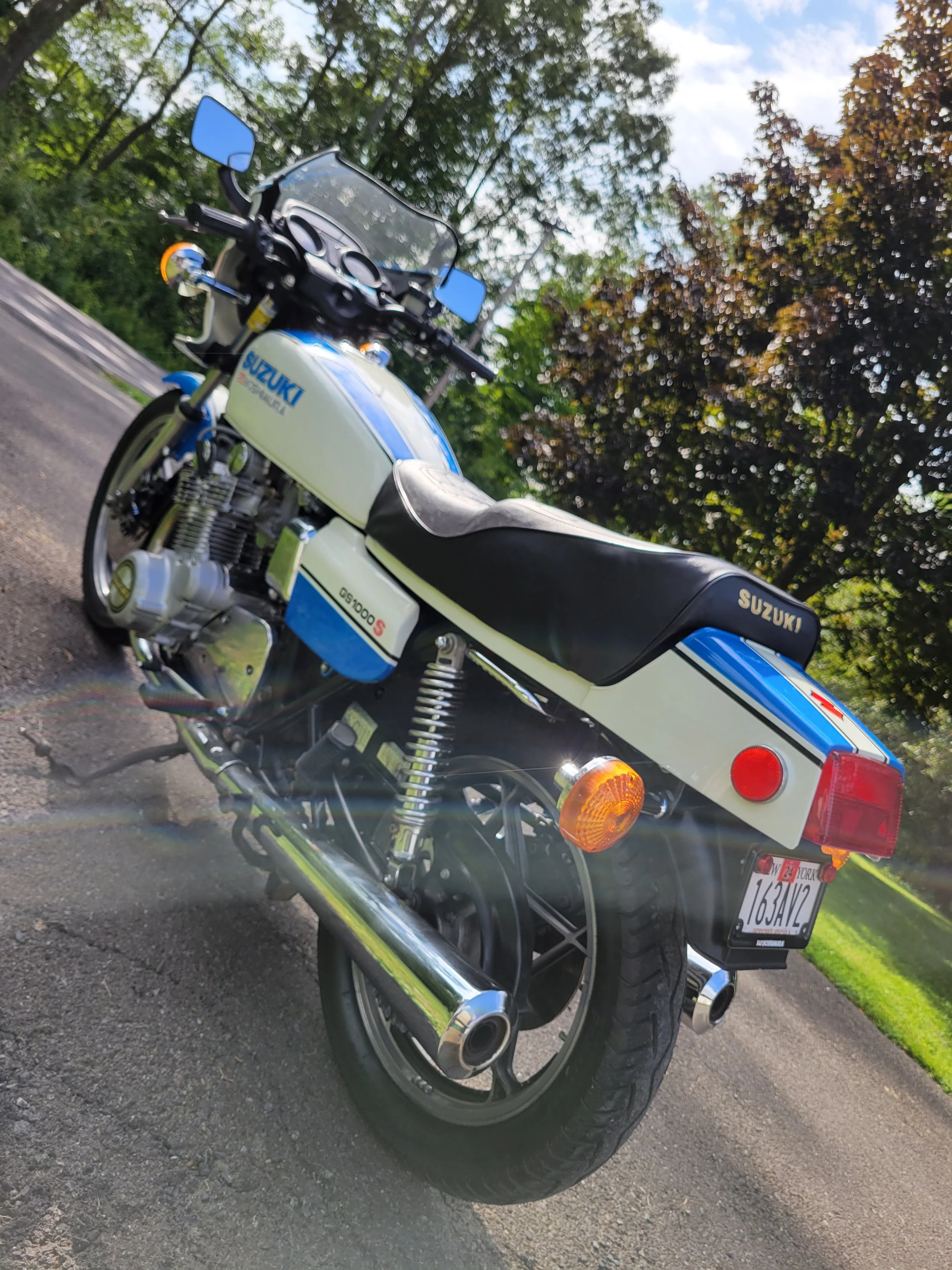 A classic Suzuki  motorcycle parked on a paved road, with trees and a partly cloudy sky in the background.