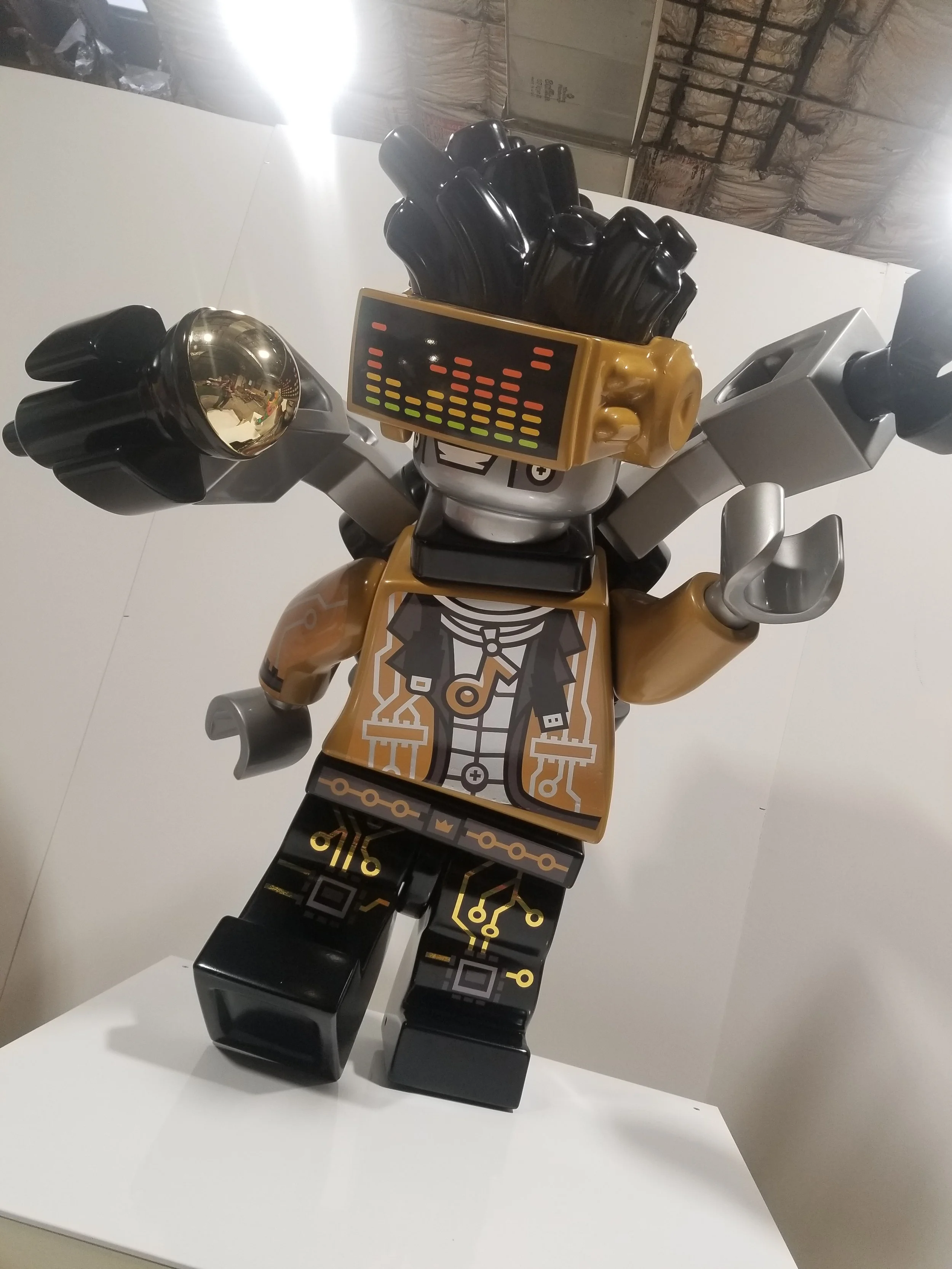 A robot sculpture made of LEGO bricks, with electronic circuit patterns and a digital sound wave display on its head, standing on a white surface.