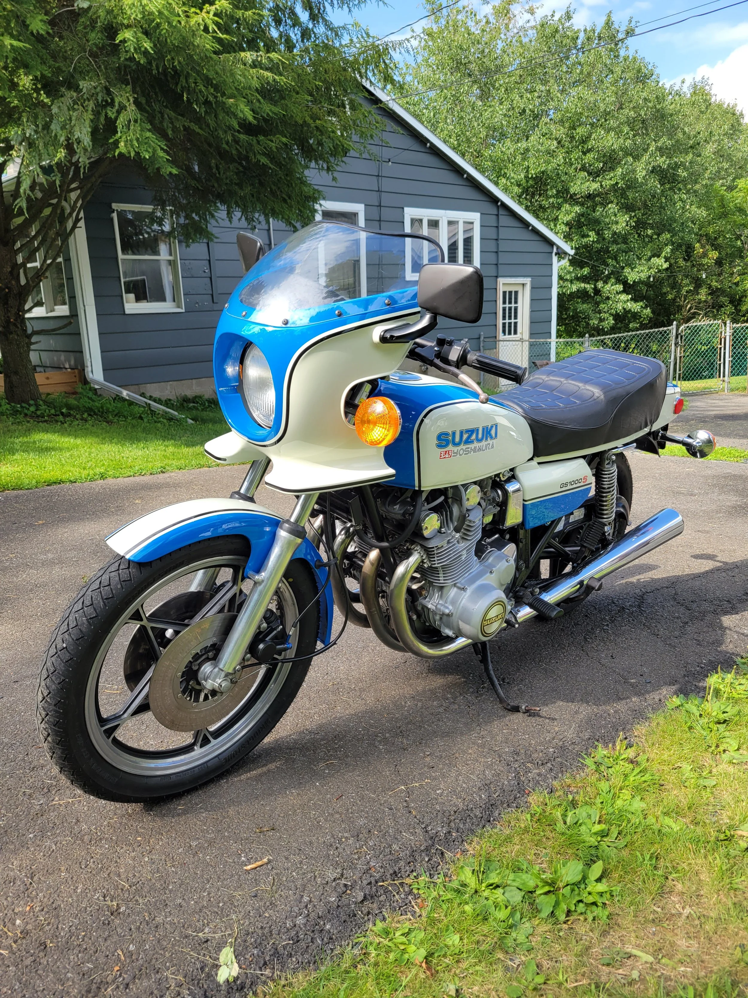 A vintage Suzuki motorcycle parked on a driveway with a blue and white color scheme, featuring a front fairing with a windshield, black seat, and chrome exhaust pipes, with a house and trees in the background.