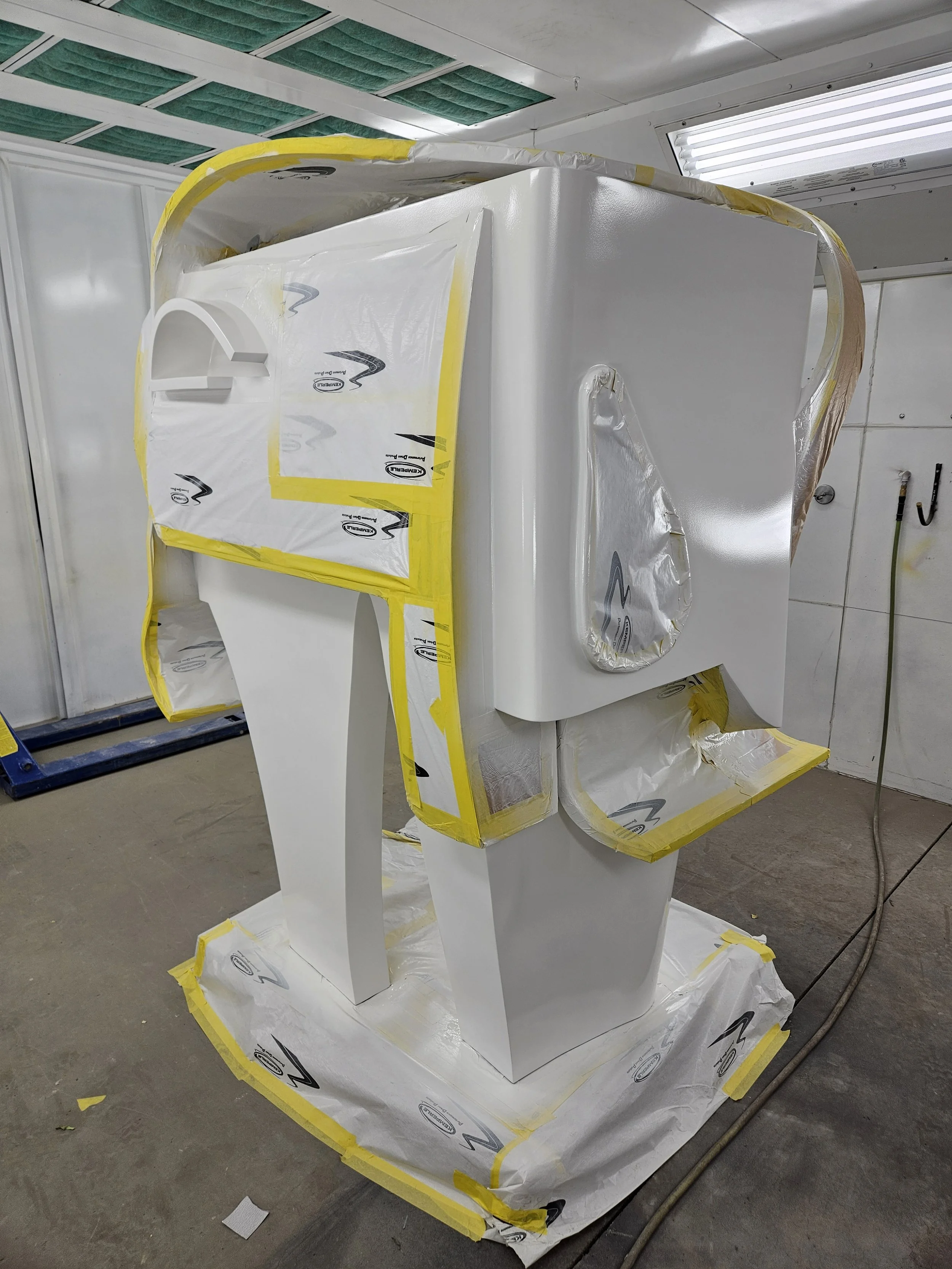 A white car hood being painted in a paint booth, covered with protective paper and tape, with the surrounding area cleaned and prepared for painting.