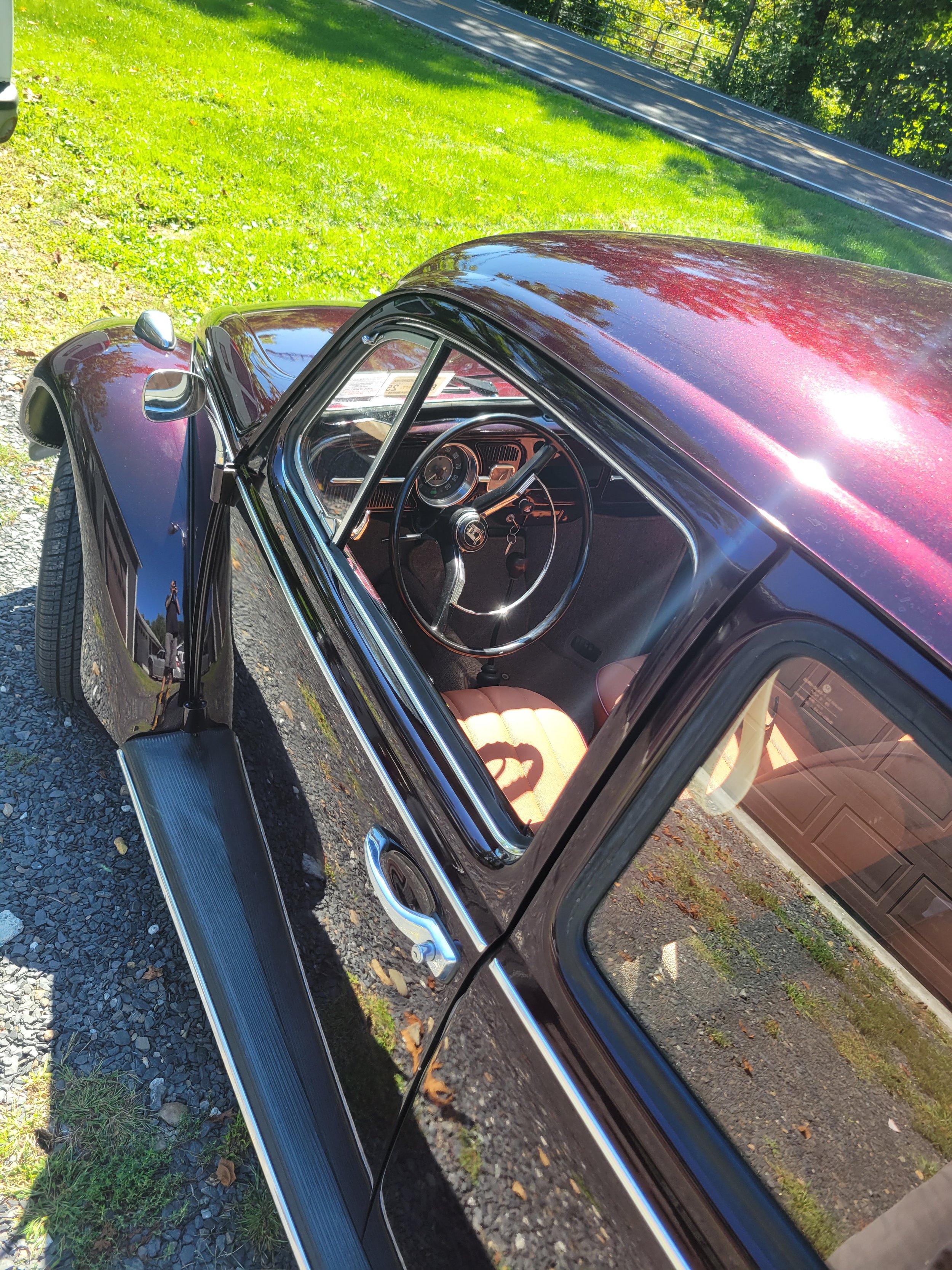 A vintage black and red car parked on a grassy area next to a road, showing the interior steering wheel and dashboard through the window.
