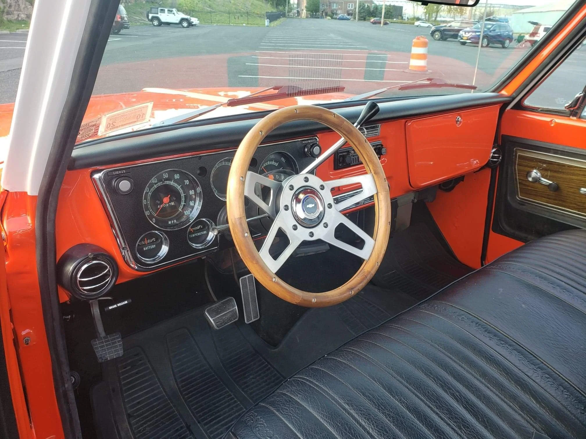 The interior of a vintage orange Chevrolet truck showing a black dashboard, a wooden steering wheel, and black leather seats. The dashboard has classic gauges including speedometer, gas, oil, and battery meters.