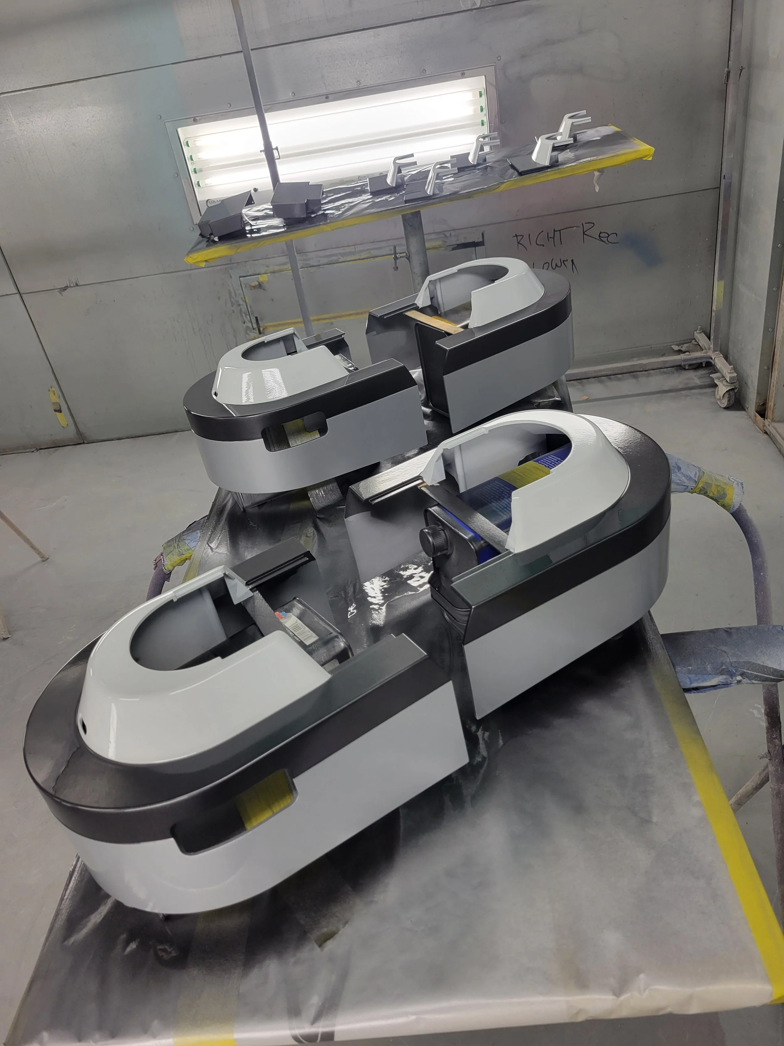 Four white and black futuristic-looking machines with open slots sit on a metallic work surface. In the background, an illuminated fluorescent light fixture is mounted on a gray wall with handwritten text that reads 'RIGHT Rod Down.'