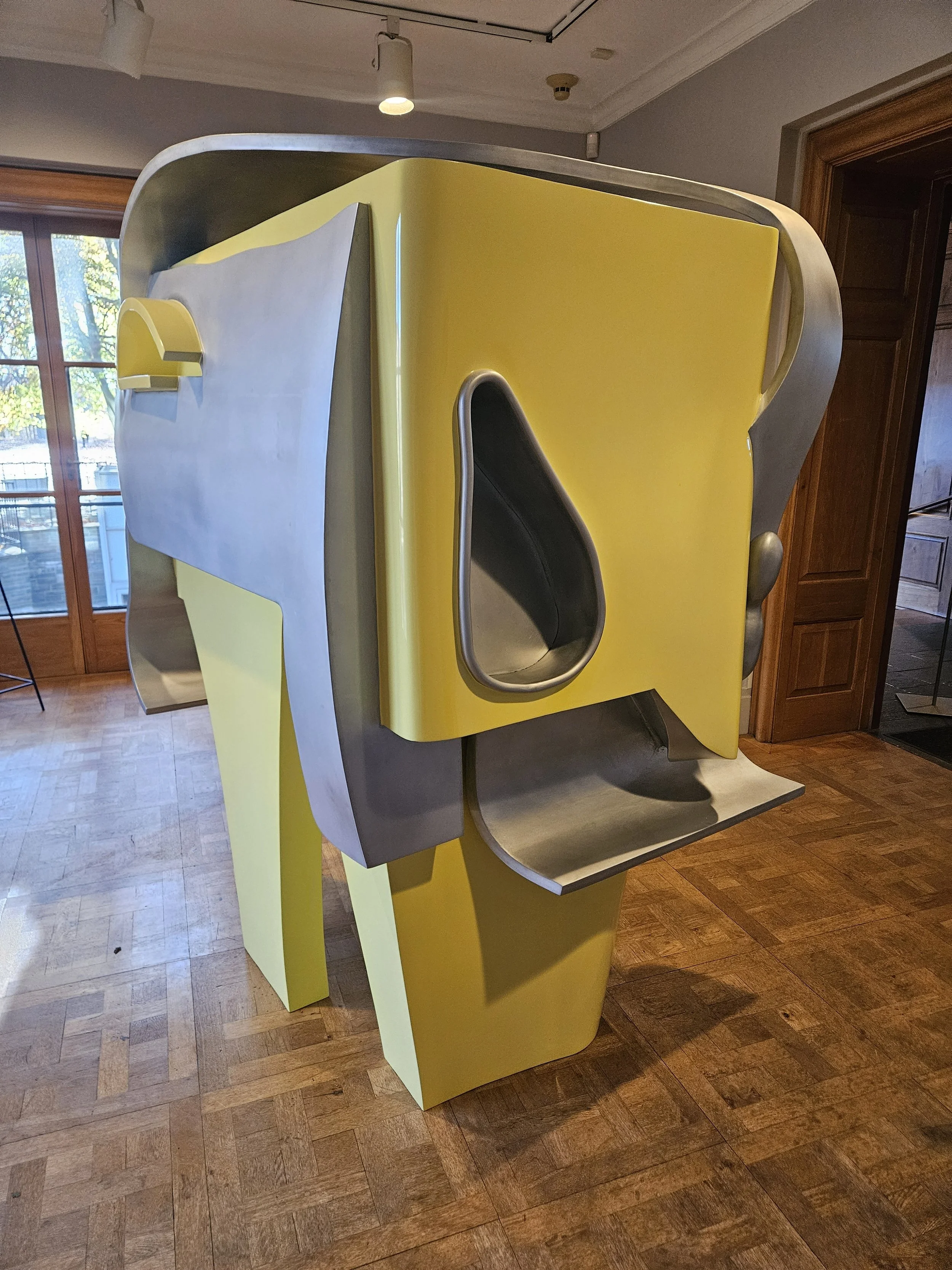 Contemporary art sculpture with a yellow and gray color scheme, featuring geometric shapes and hollowed sections, placed indoors on wooden flooring near a window.