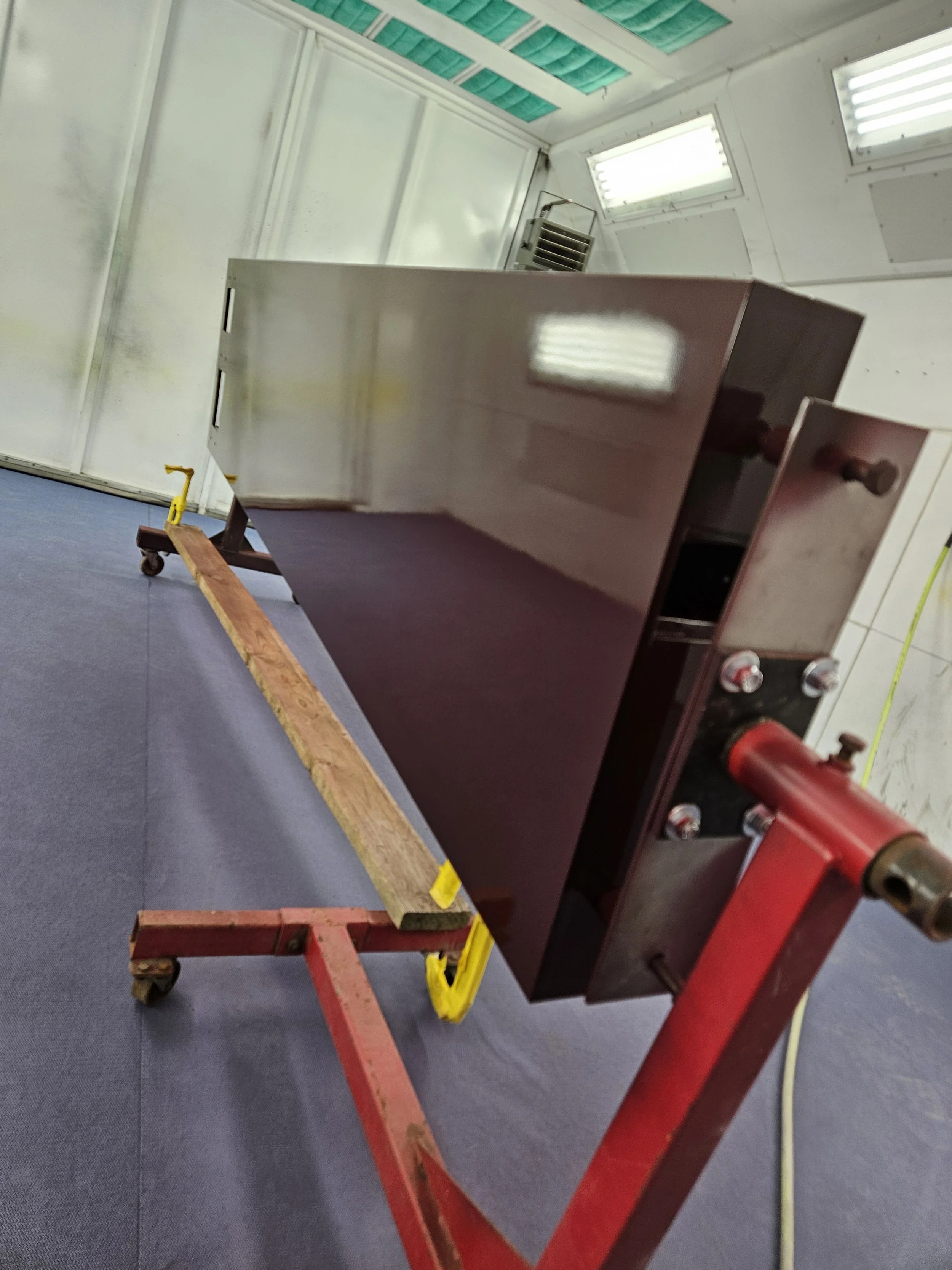 A stainless steel appliance, possibly a mini fridge or microwave oven, is secured on a red and yellow adjustable cart with wheels in a workshop or laboratory setting. The background features white walls, several windows, and vents.
