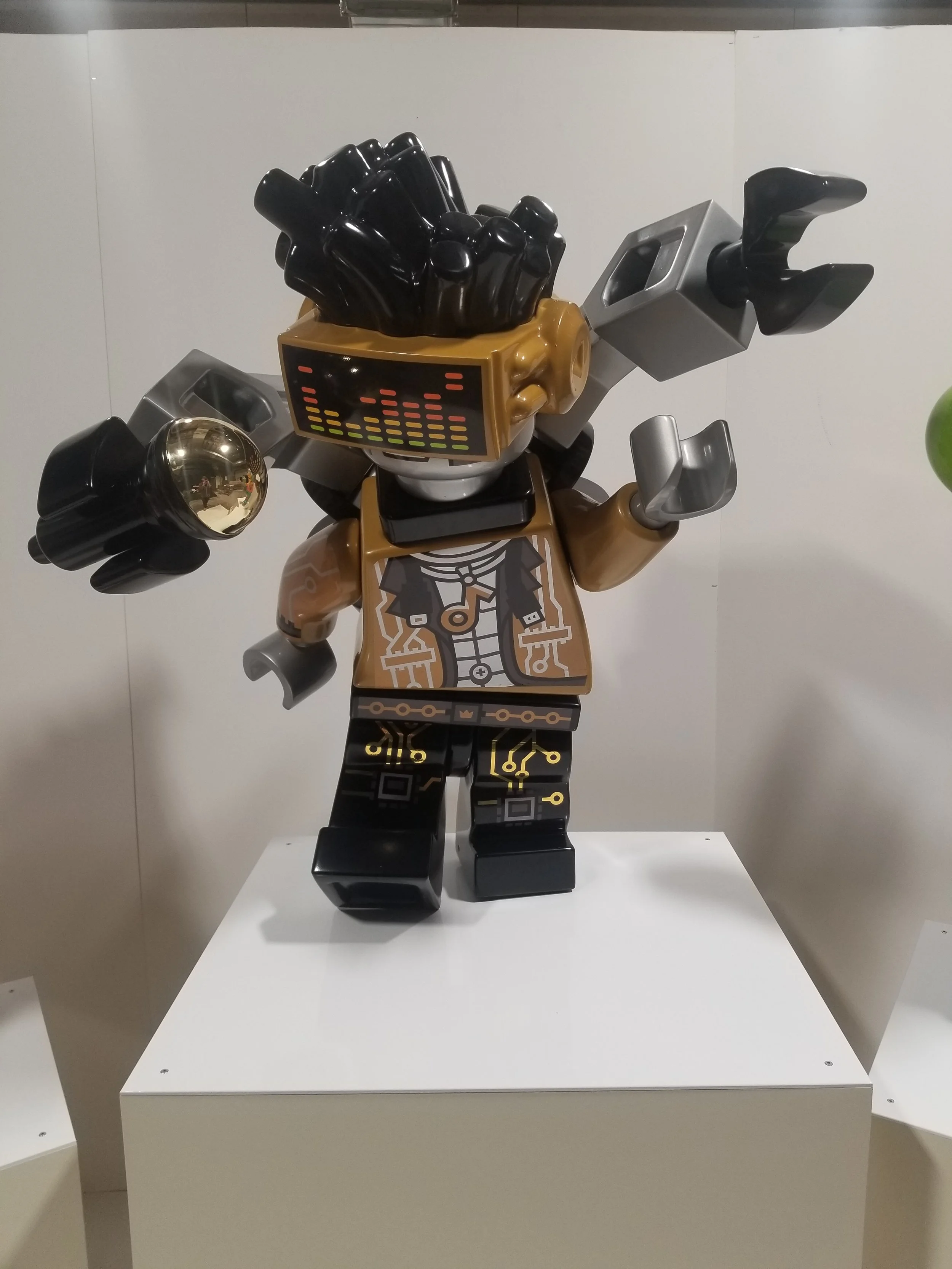 A LEGO robot sculpture with a square head featuring a light display, black and gold hair, and headphones. It has a tan jacket, printed shirt, and circuit-like designs on its black legs.