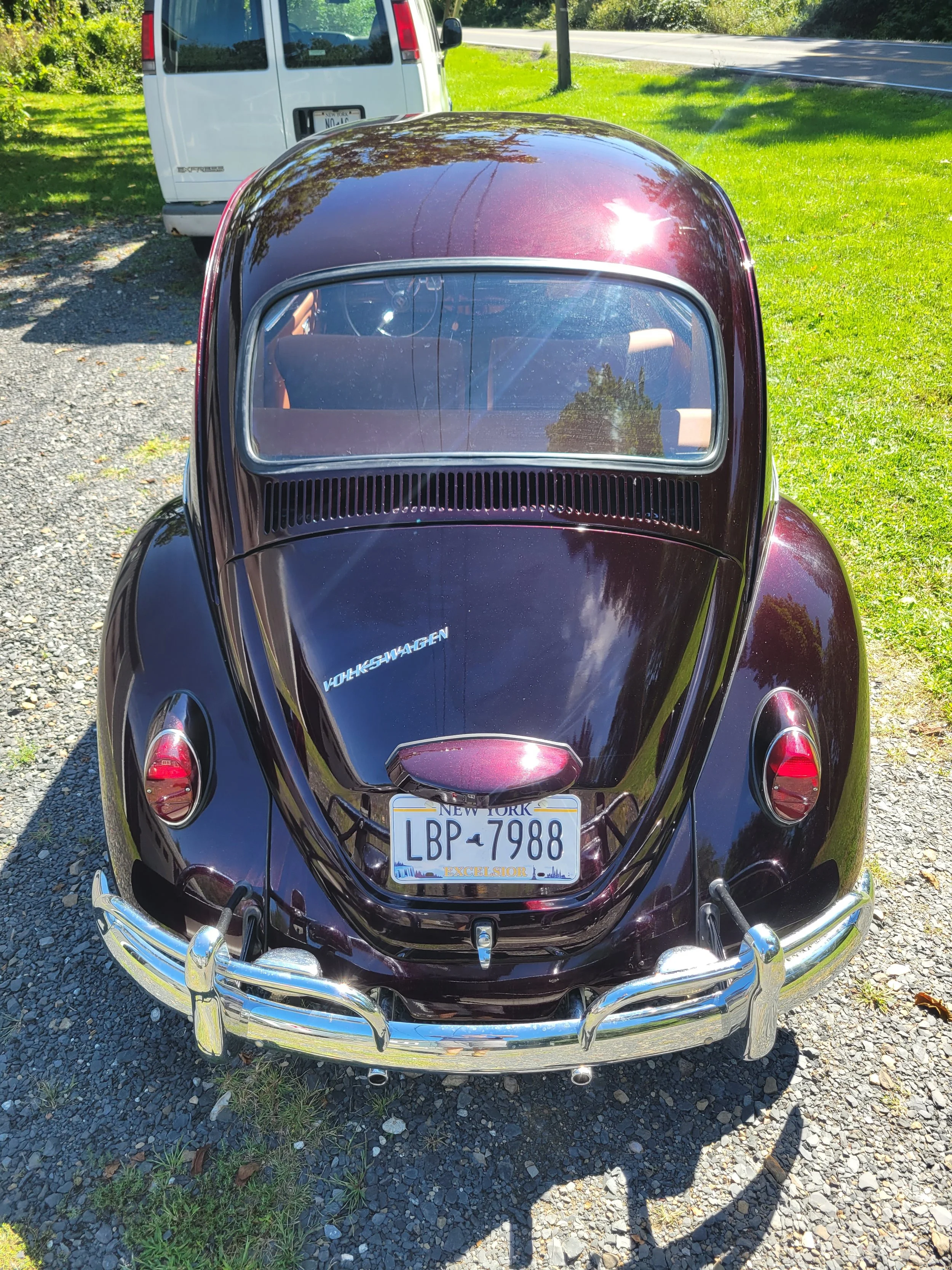 Rear view of a vintage black Volkswagen Beetle with a New York license plate, parked on a gravel area next to a grassy field and a white van. The car has chrome bumpers and a curved rear window.