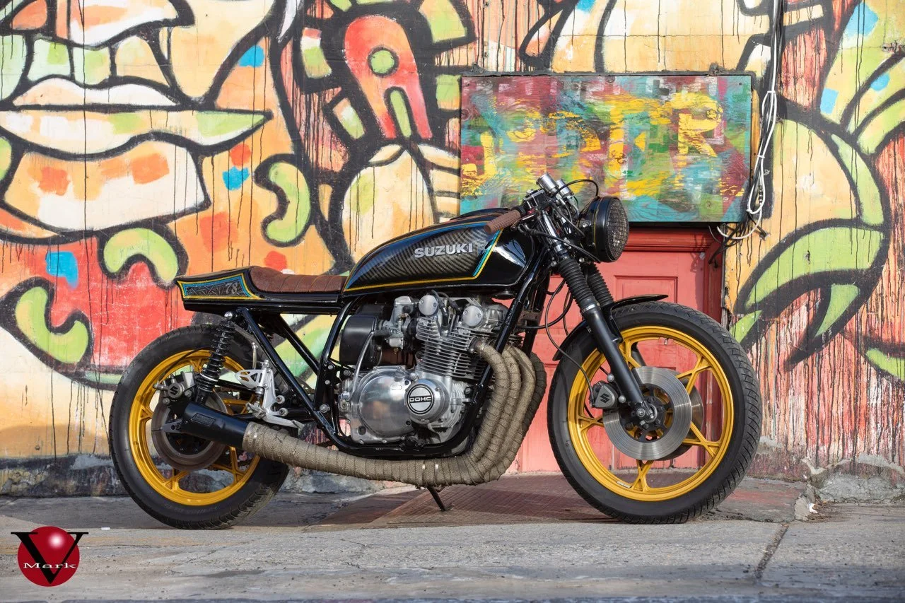 A custom Suzuki motorcycle with black body and yellow wheels parked in front of a colorful graffiti-covered wall.