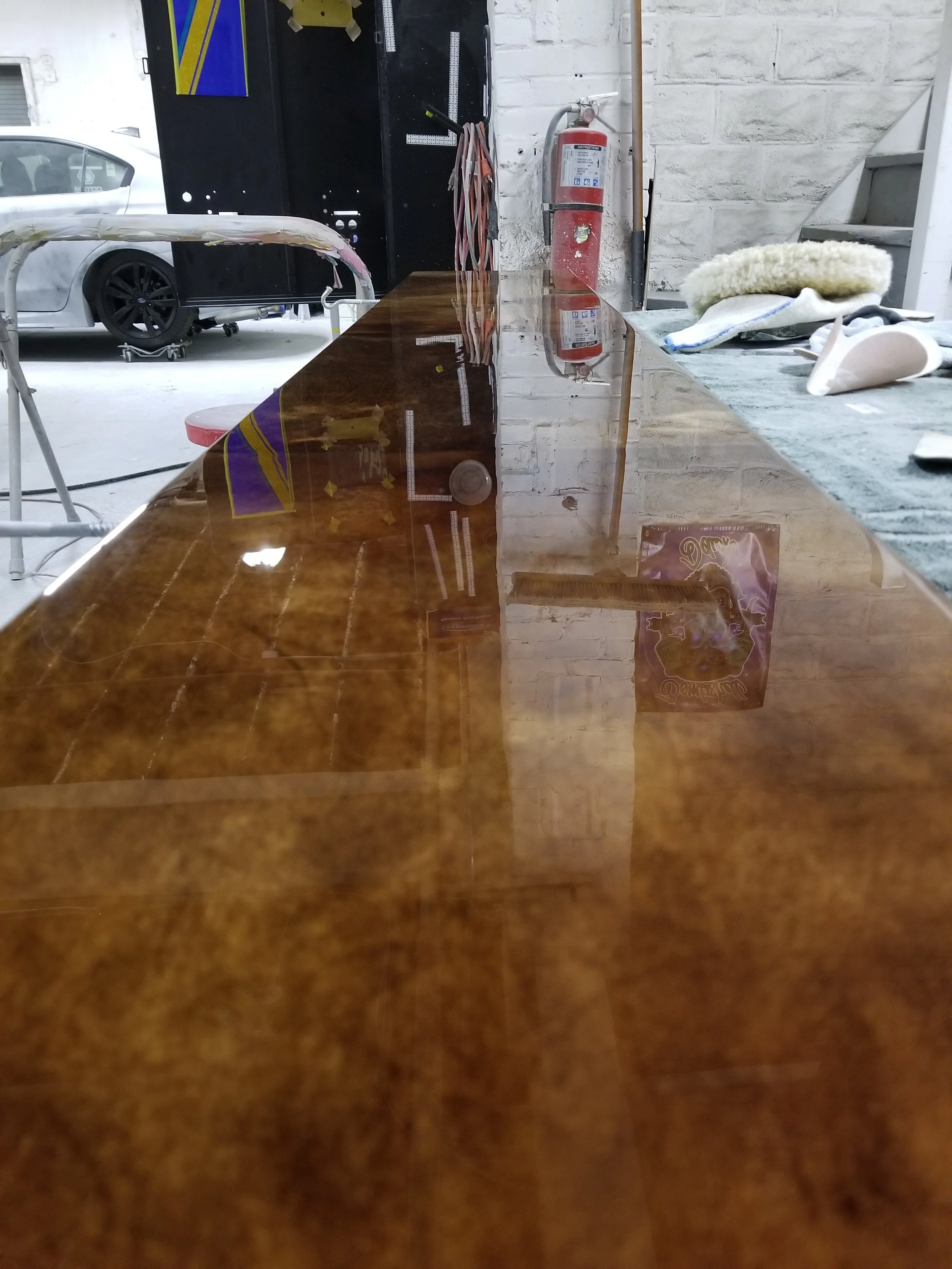 A polished wooden table with reflections of objects and posters in an indoor workshop or garage space. Behind the table are cleaning supplies and a fire extinguisher, with a white brick wall and some equipment around.