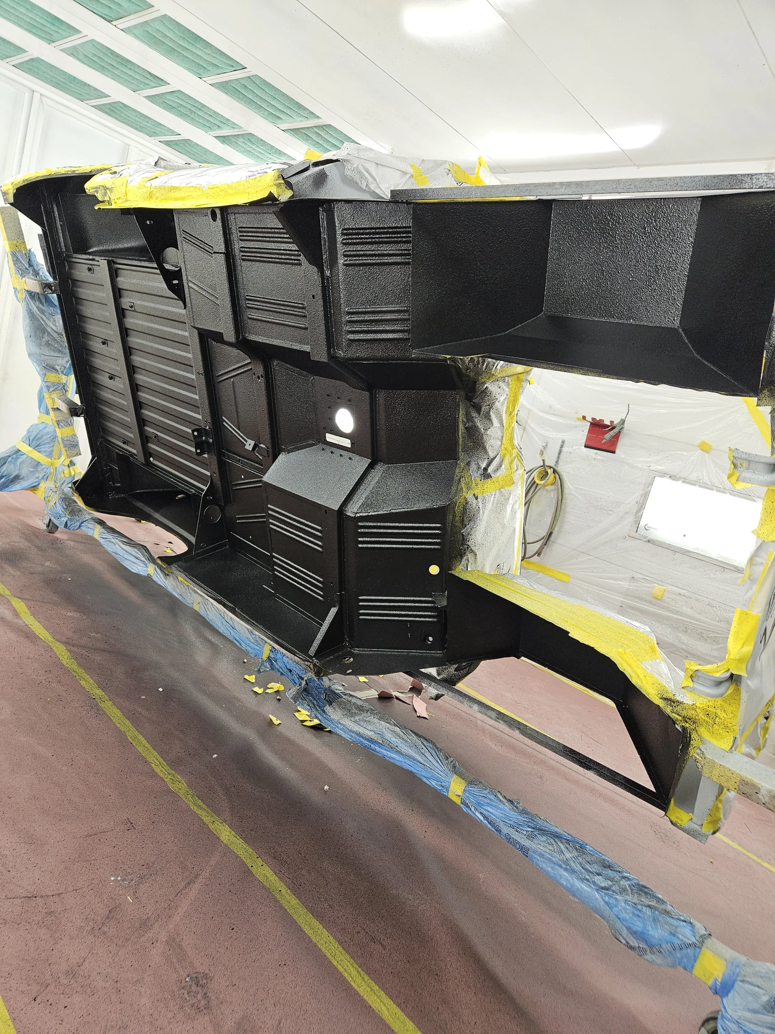 The underside of a black vehicle's interior panel is shown, surrounded by protective and masking materials in a workshop or paint booth.