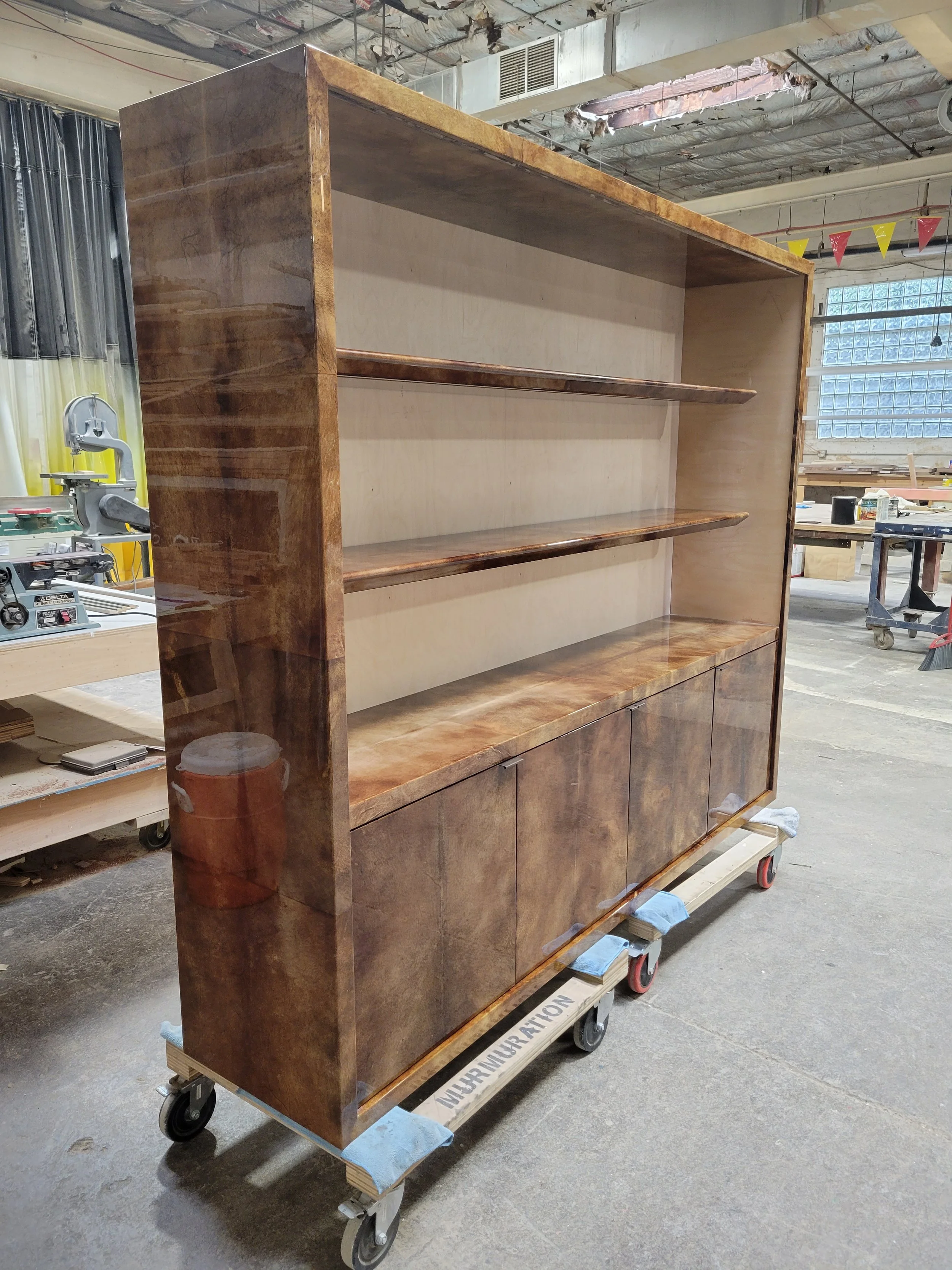A large wooden cabinet with multiple shelves and closed storage compartments, on a wheeled platform in a woodworking shop.