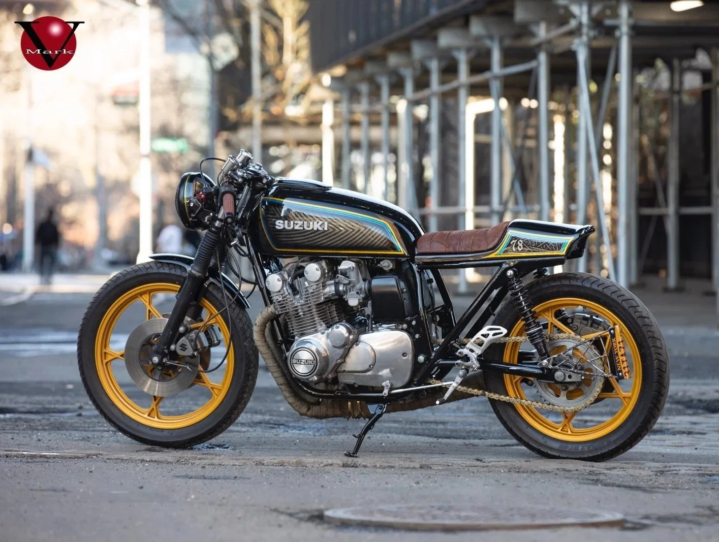A custom black and gold vintage Suzuki motorcycle with a brown leather seat parked on a city street with scaffolding in the background.